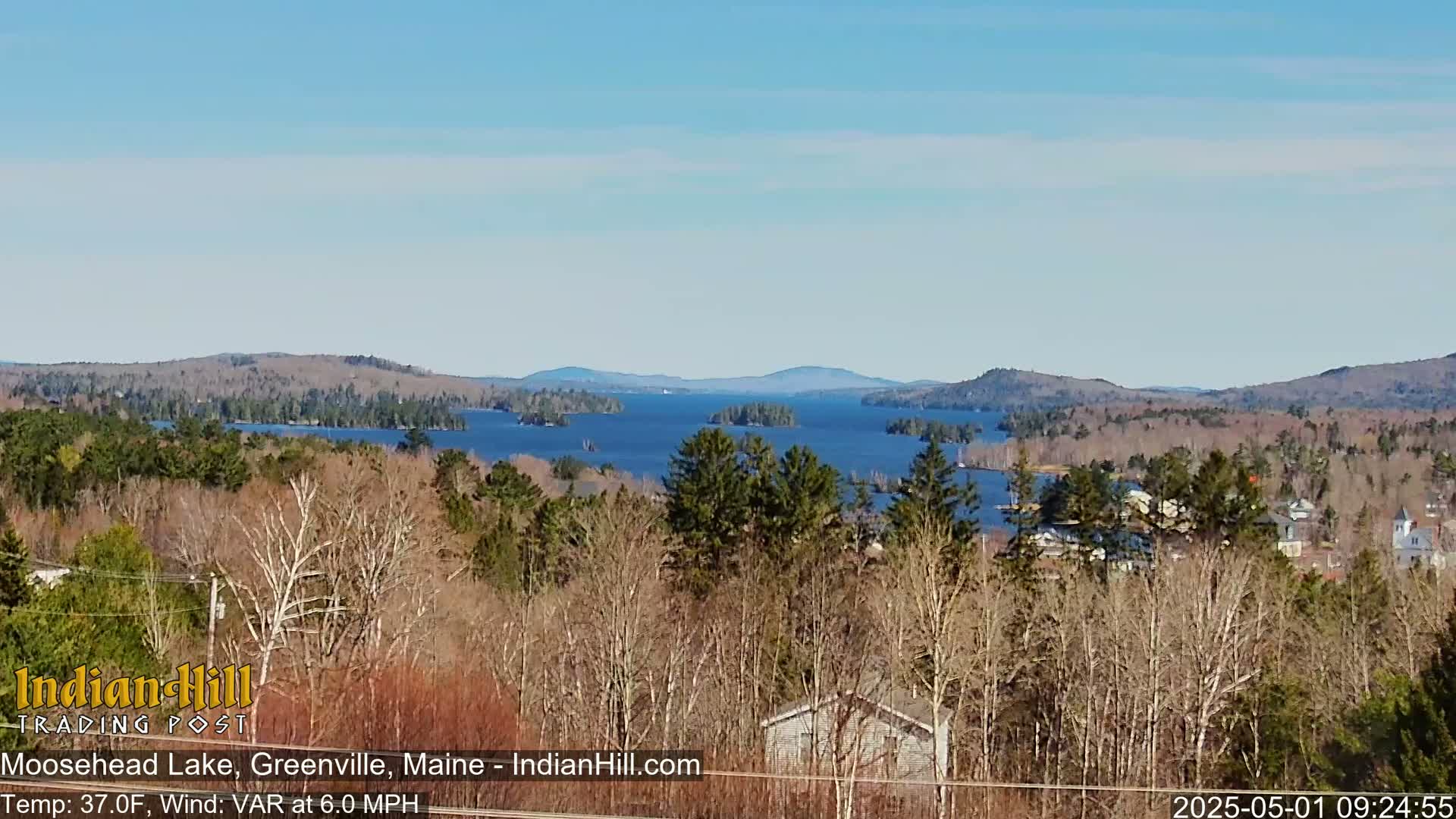 A panoramic view of Moosehead Lake, under a clear sky, shows a sparsely wooded shoreline with a few houses visible in the foreground.