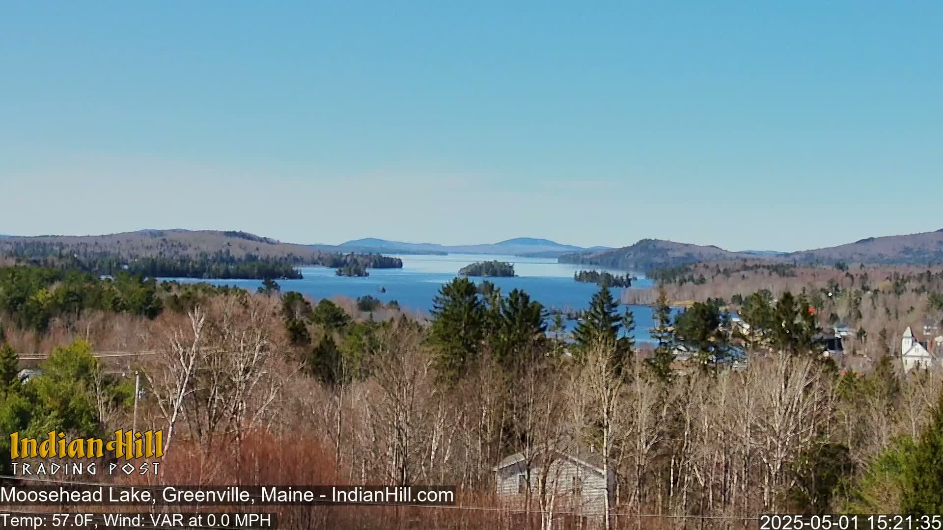 A panoramic view shows a large lake dotted with islands, surrounded by a mostly bare forest, under a clear blue sky.