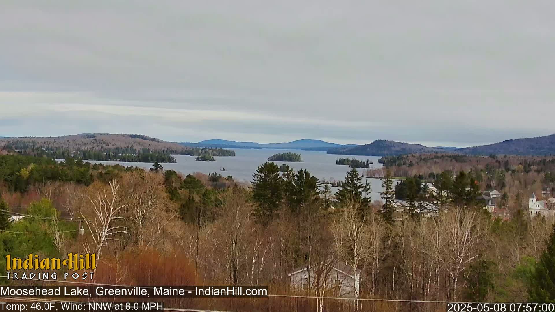 A mostly cloudy view overlooks a lake with several islands, surrounded by a forest of mostly bare trees, with mountains in the distance.