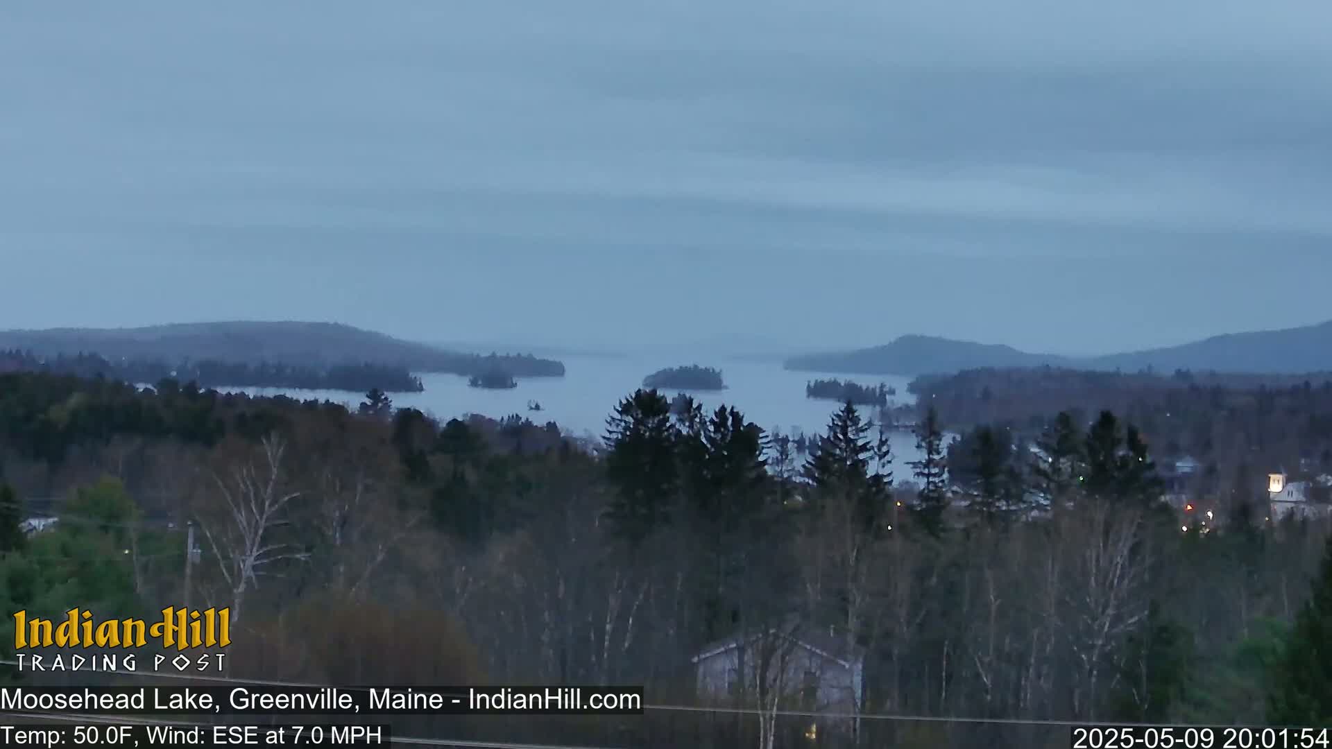 A tranquil, overcast view shows a large lake dotted with islands, surrounded by dark green forests under a dusky sky.