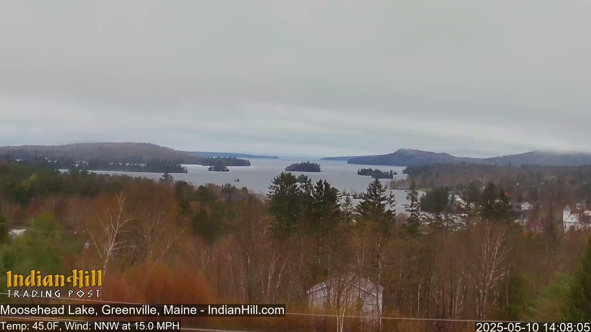 A wide, expansive view of Moosehead Lake, partially obscured by a hazy, overcast sky, shows a tranquil landscape of forested hills and numerous islands.