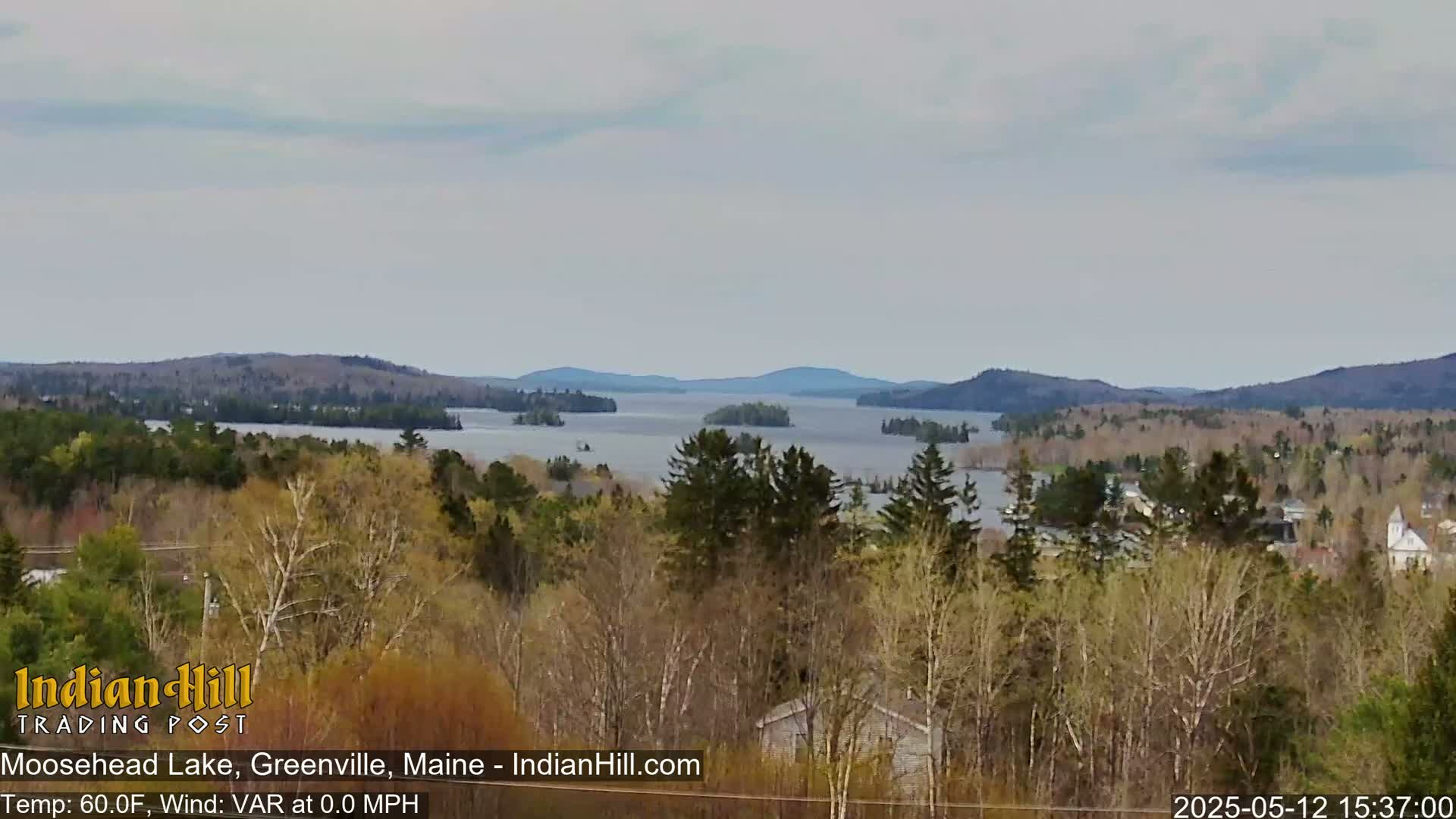A tranquil view of Moosehead Lake, under a mostly cloudy sky, shows a large body of water surrounded by forested hills and trees in varying shades of green and brown.