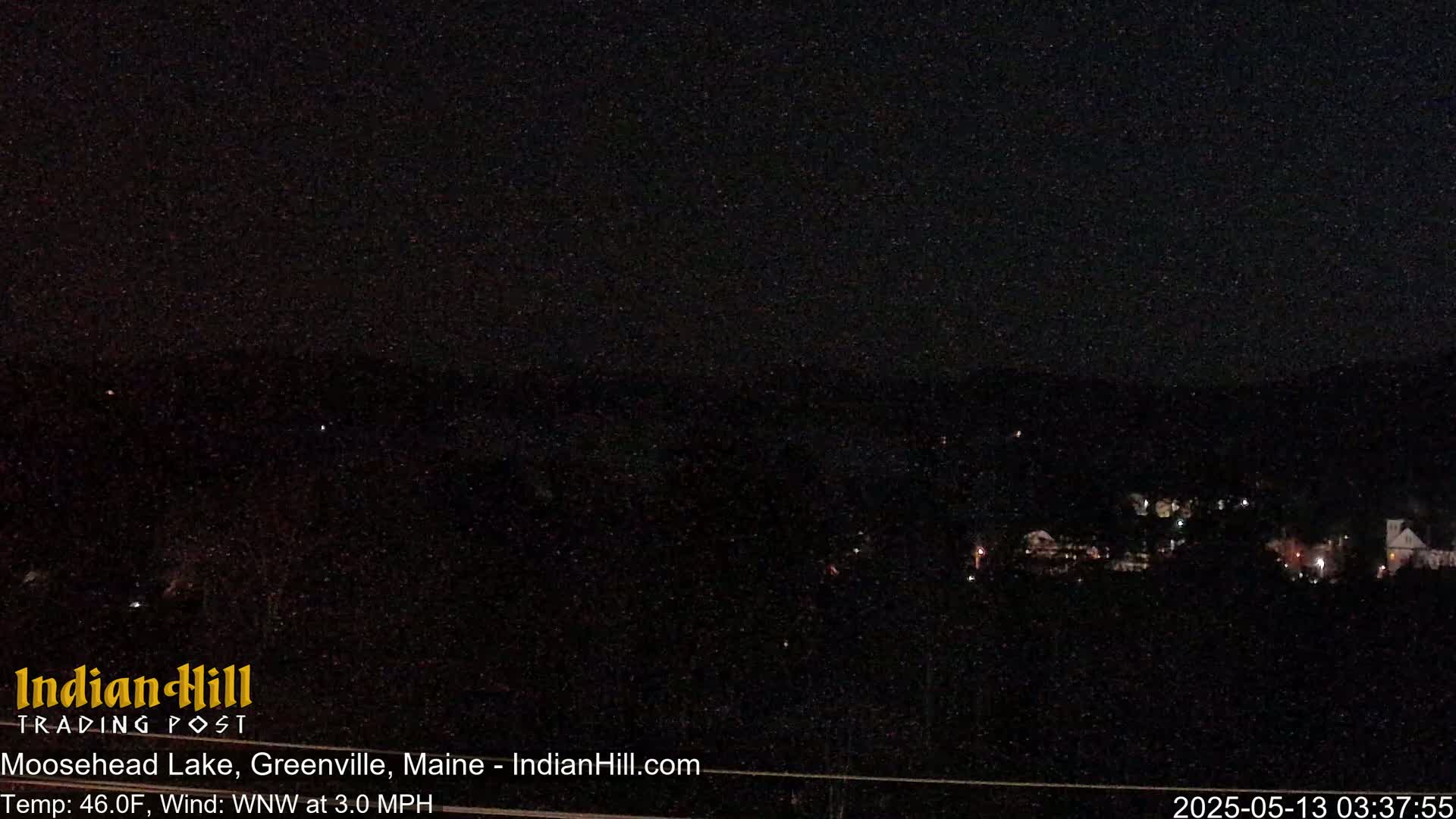 A dark, nighttime view of a town with a few lights visible, under a clear, dark sky with a light wind.