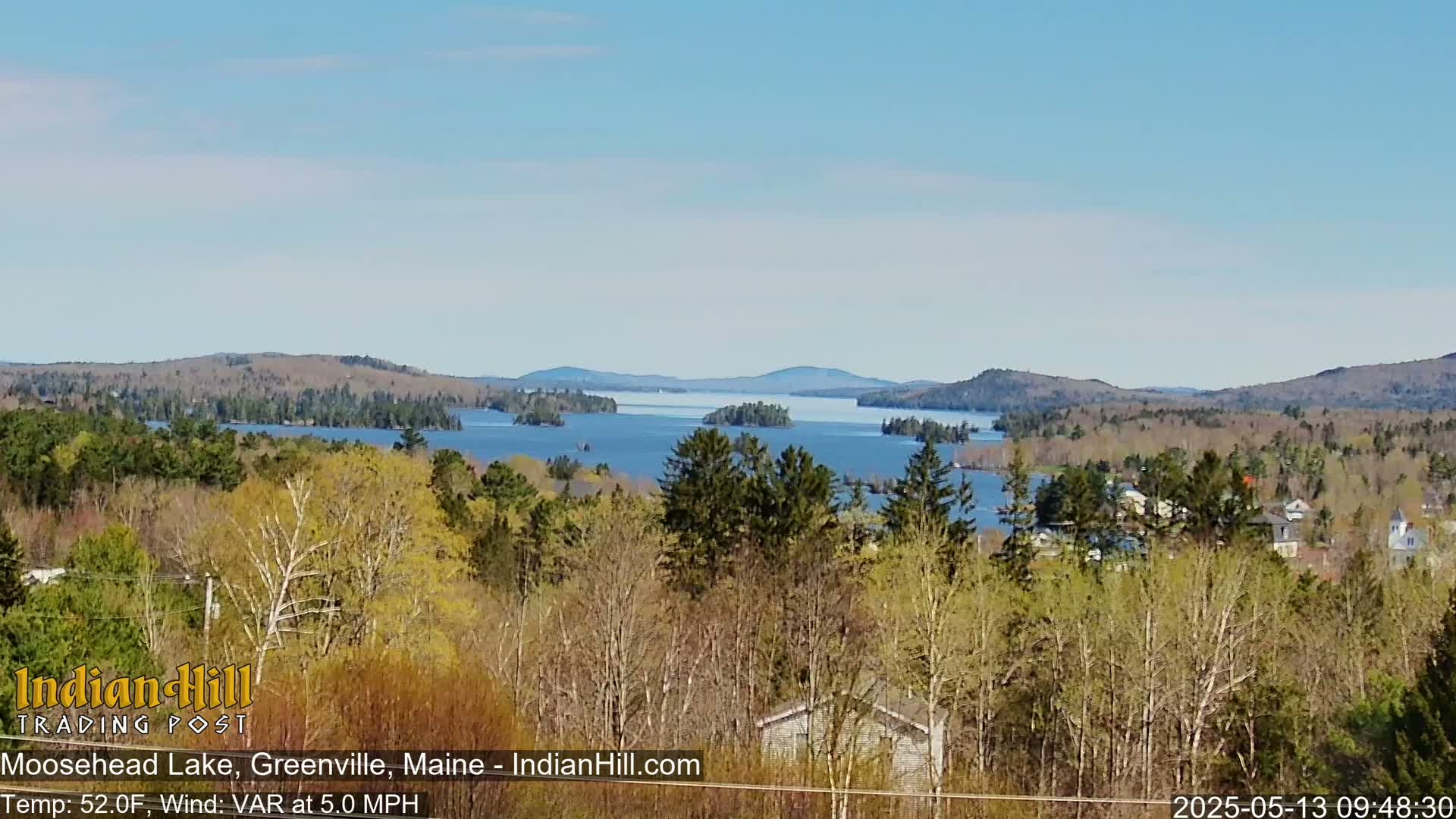 A panoramic view of a lake dotted with islands, surrounded by a mix of evergreen and deciduous trees under a clear, bright sky.