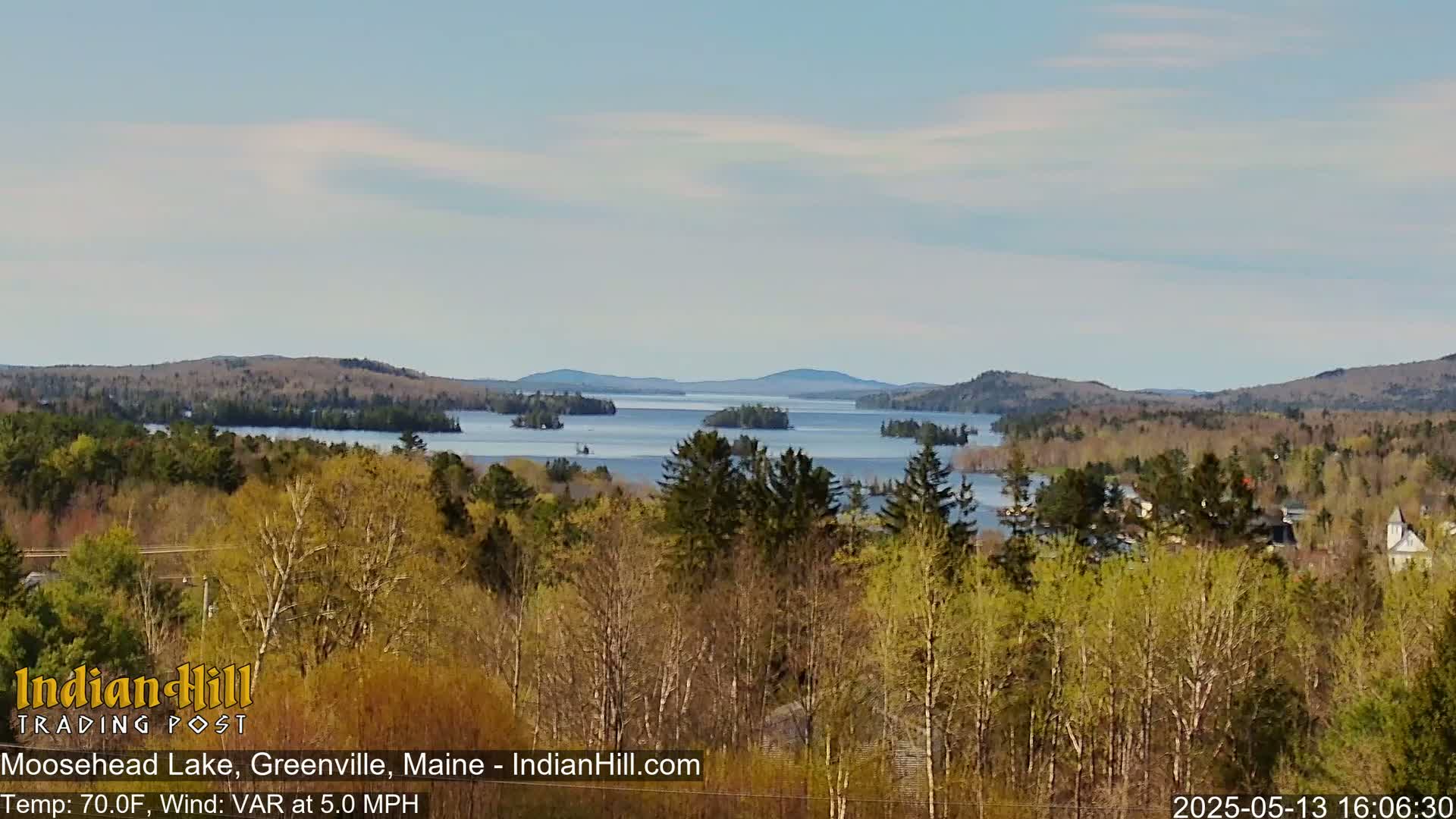 A tranquil lake dotted with islands stretches to distant mountains under a mostly clear sky, viewed from a wooded hillside.