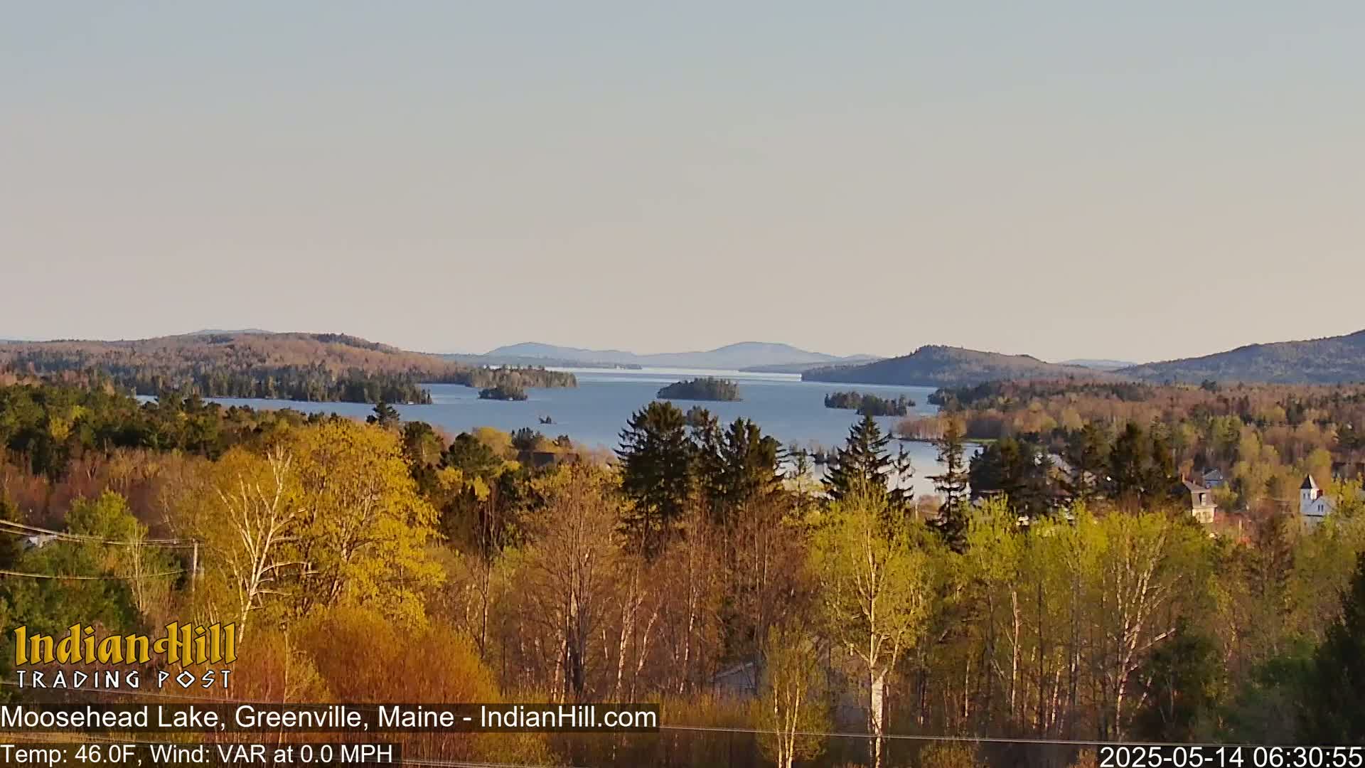 A view from a hill overlooking a large lake dotted with islands, surrounded by trees showing early spring foliage under a clear, light sky.
