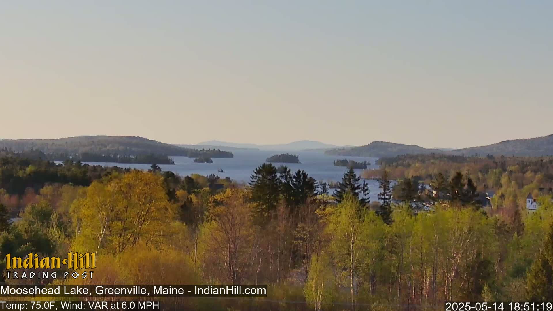 A tranquil lake dotted with islands is seen from an elevated vantage point, surrounded by lush, spring-colored forests under a clear, light sky.