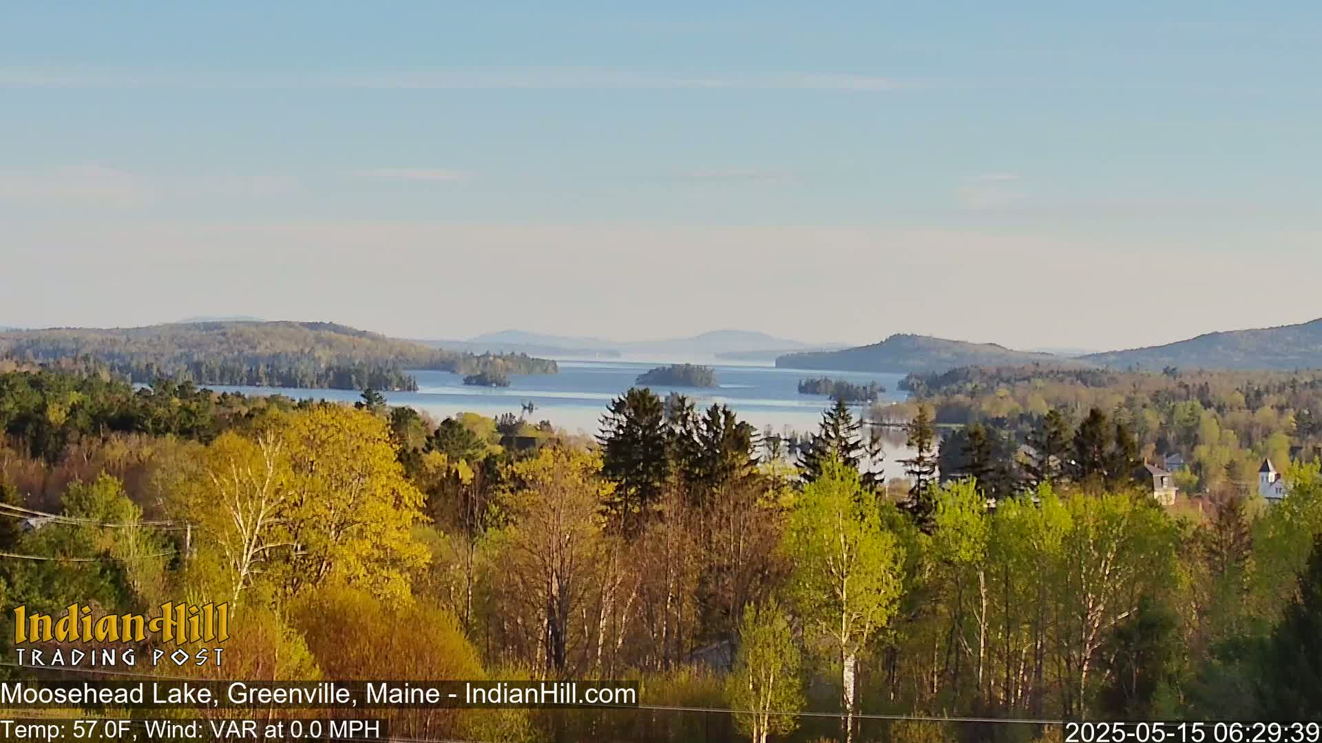 A calm, clear day reveals a view of Moosehead Lake from a wooded hillside, with the water partially obscured by a light fog.