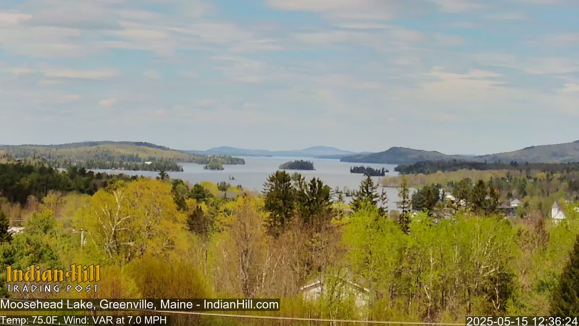 A tranquil lake dotted with islands is surrounded by a lush, green forest under a partly cloudy sky.