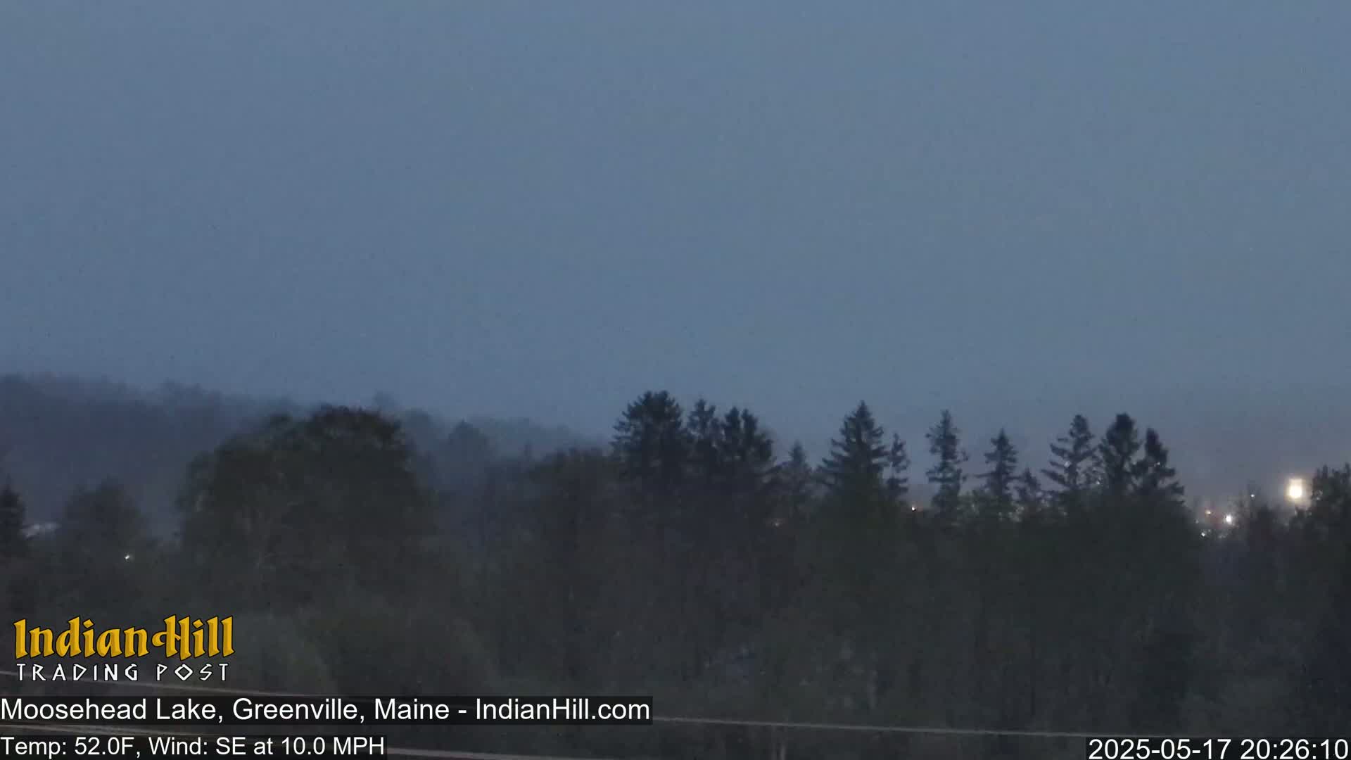 A hazy, dark-blue evening sky over a line of dark evergreen trees under a southeast wind.