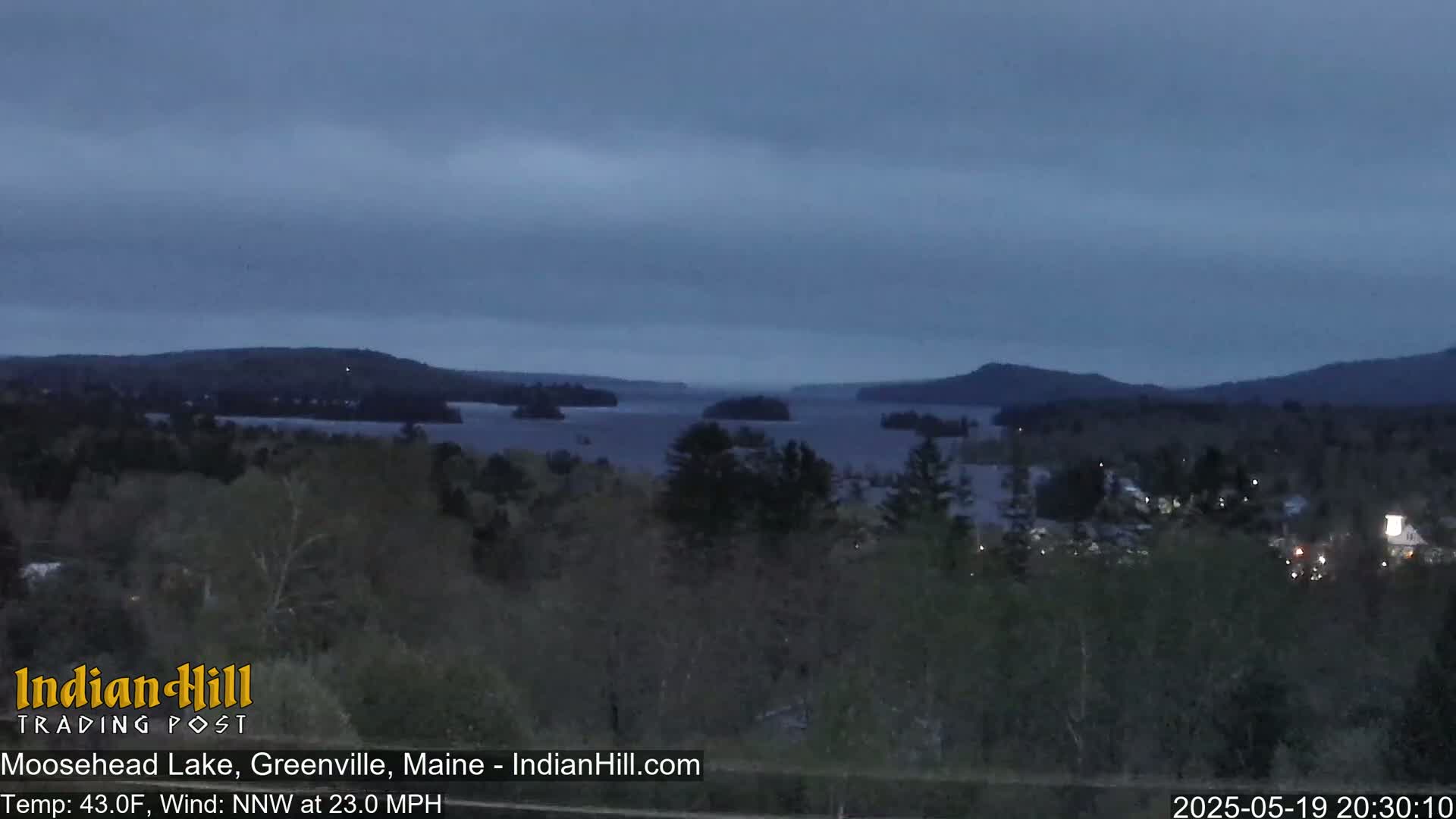 A dark, overcast evening scene shows a large lake with several islands, surrounded by dark green forested hills.