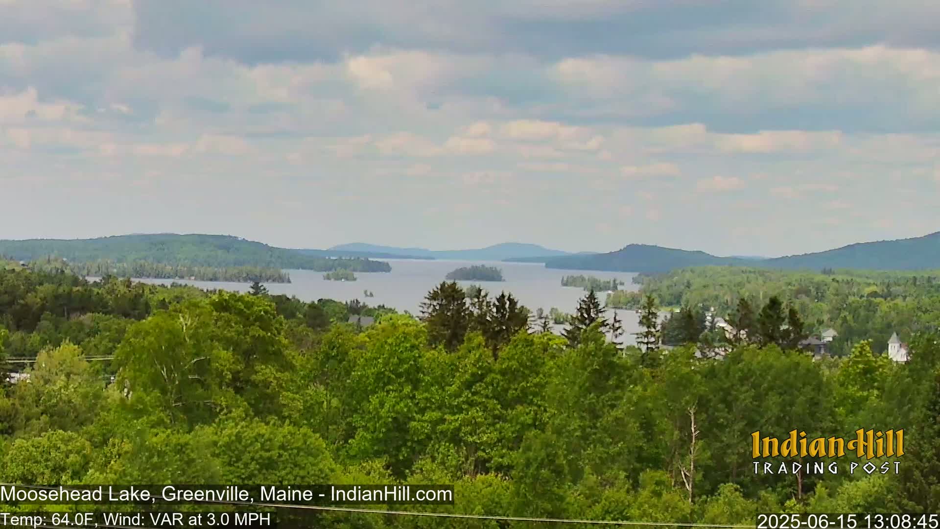 A view of a large lake surrounded by lush green forests and mountains under a partly cloudy sky.