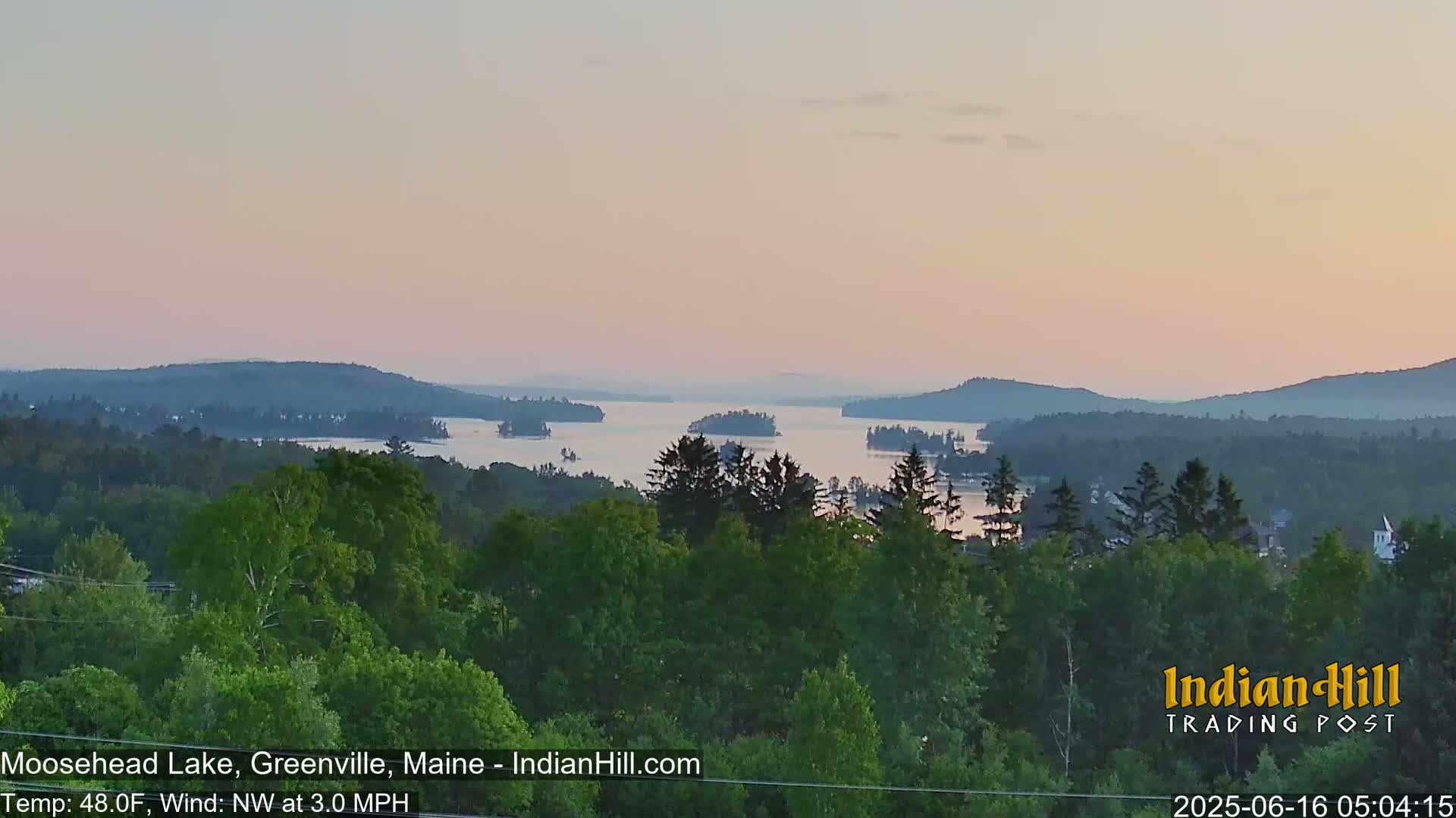 A calm, pinkish sunrise illuminates a lake with several islands, viewed from a forested hill.