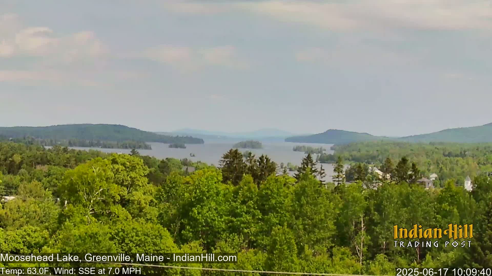 A view of a large lake surrounded by lush green trees and mountains under a mostly cloudy sky with a gentle south-southeast wind.