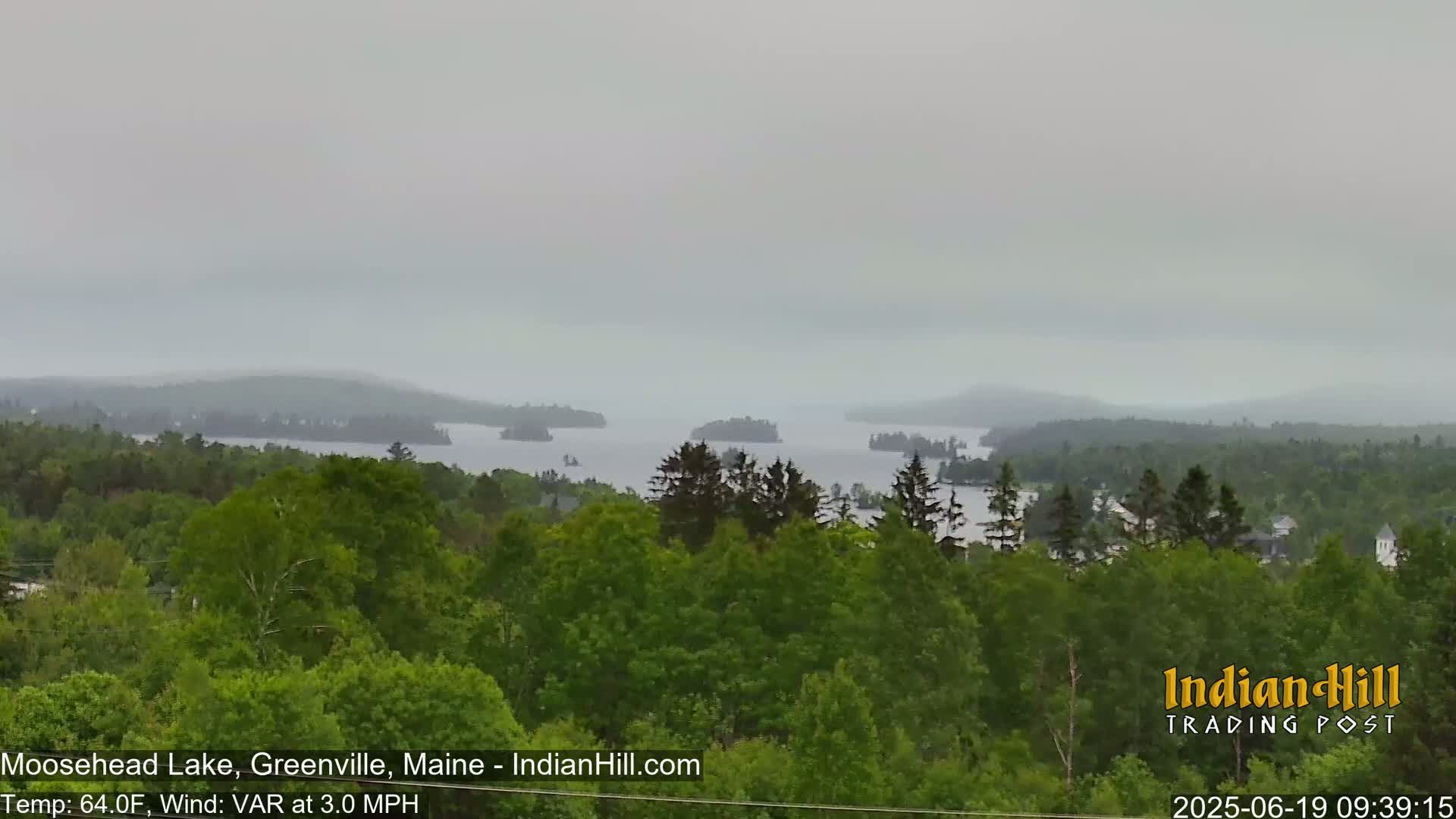 A hazy, overcast view shows a lake surrounded by lush green forests and small islands, with a slight wind.