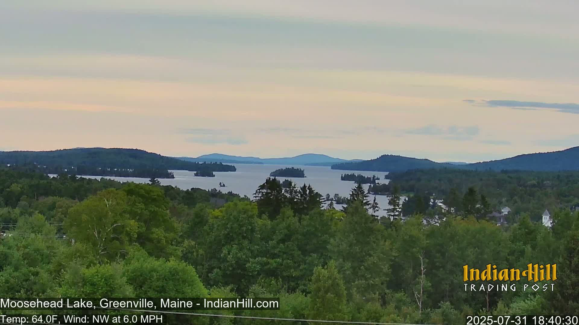 A tranquil lake dotted with islands is seen from a forested hillside under a pale, overcast sky.