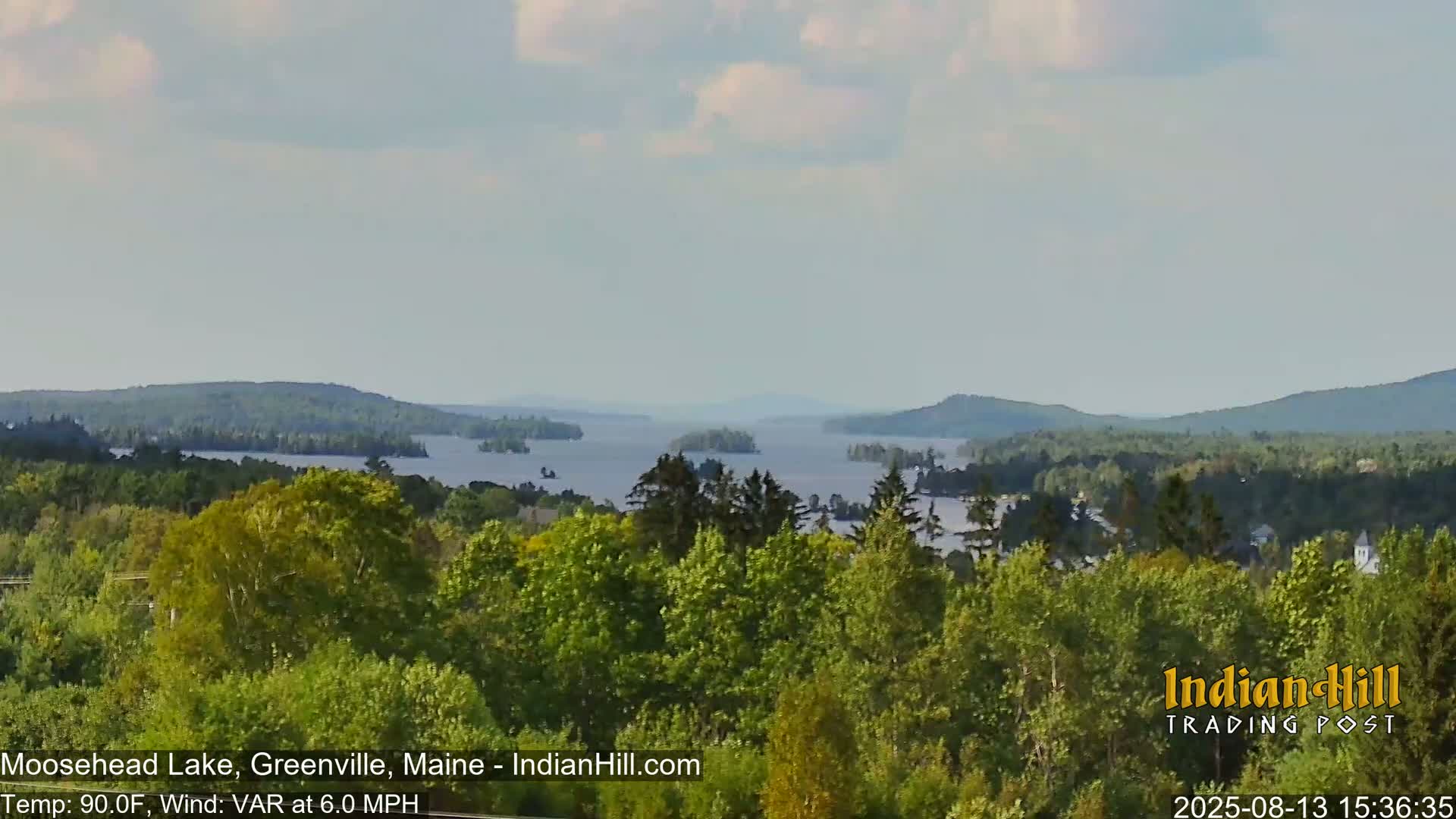 A tranquil view of a large lake dotted with islands, surrounded by lush green forests under a partly cloudy sky.
