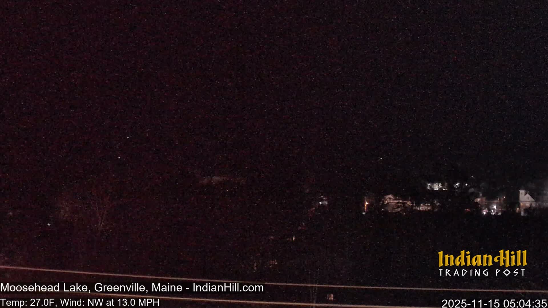 A very dark, grainy night view reveals distant scattered lights of a settlement against a dark sky, with a temperature of 27.0F and a northwest wind at 13.0 MPH.