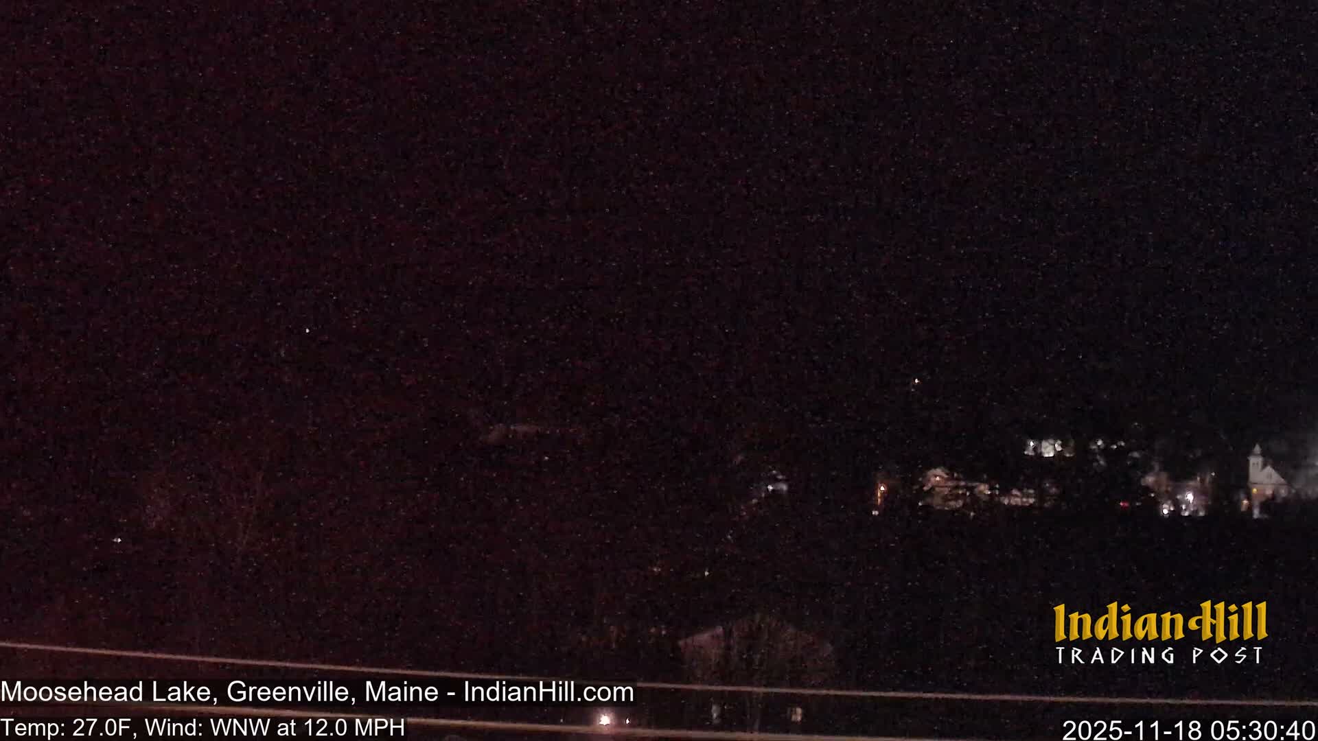 A dark and hazy outdoor scene at night reveals the silhouettes of a dense line of evergreen trees stretching across the midground under an overcast, reddish-brown sky, with a few faint lights barely visible in the distance.