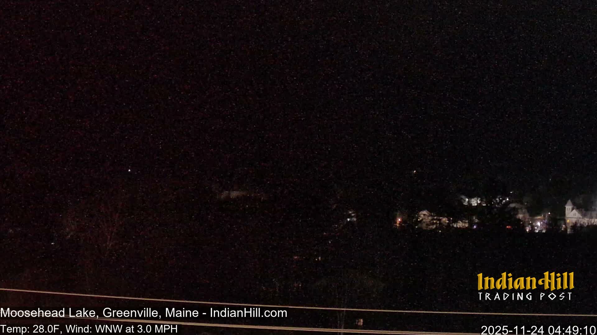 A dark nighttime scene shows a distant town with illuminated buildings and a church steeple, while faint bare trees are visible in the foreground, all under a sky filled with falling snow.