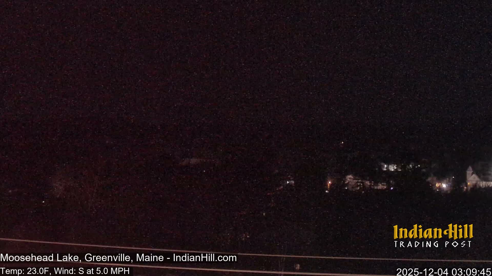 A very dark outdoor night scene reveals numerous tiny white specks, likely light snow, across the frame, with the faint glow of distant buildings and lights visible on the horizon.