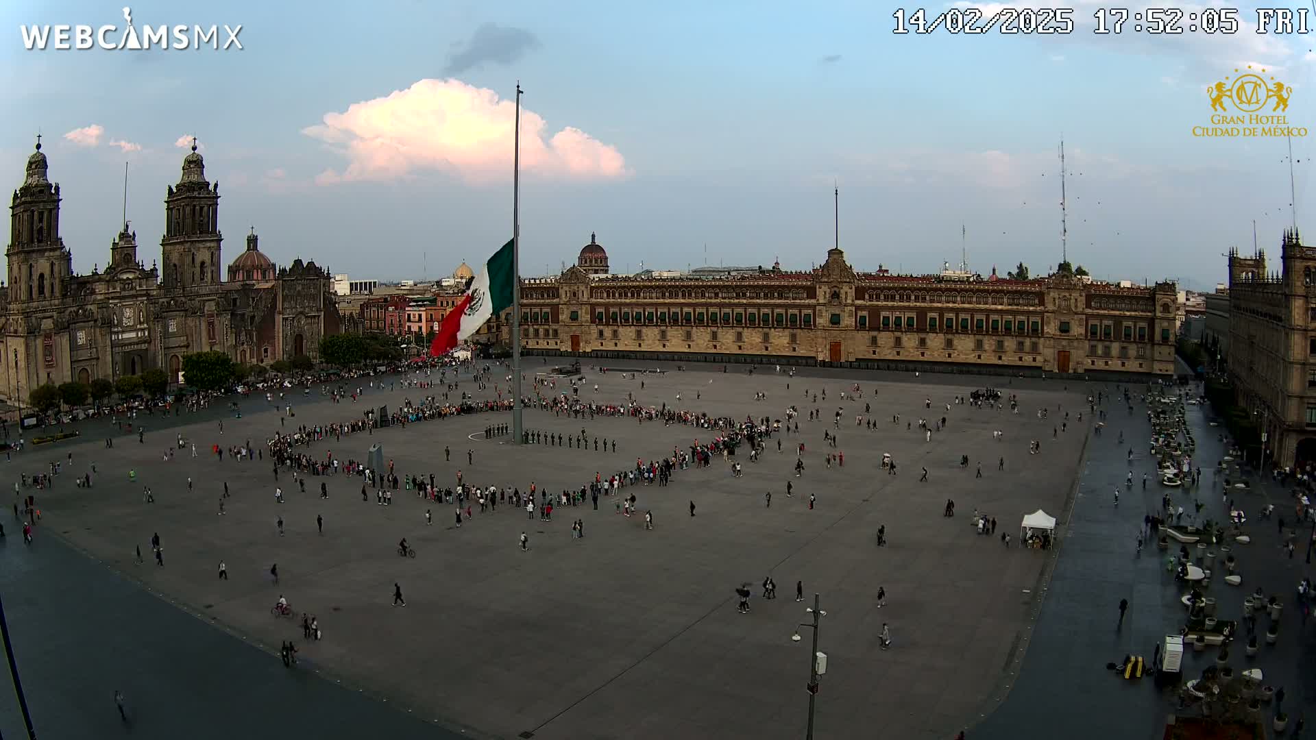 A large plaza with many people, the Mexican flag hanging from a tall pole, and a large colonial-style building under a partly cloudy sky.