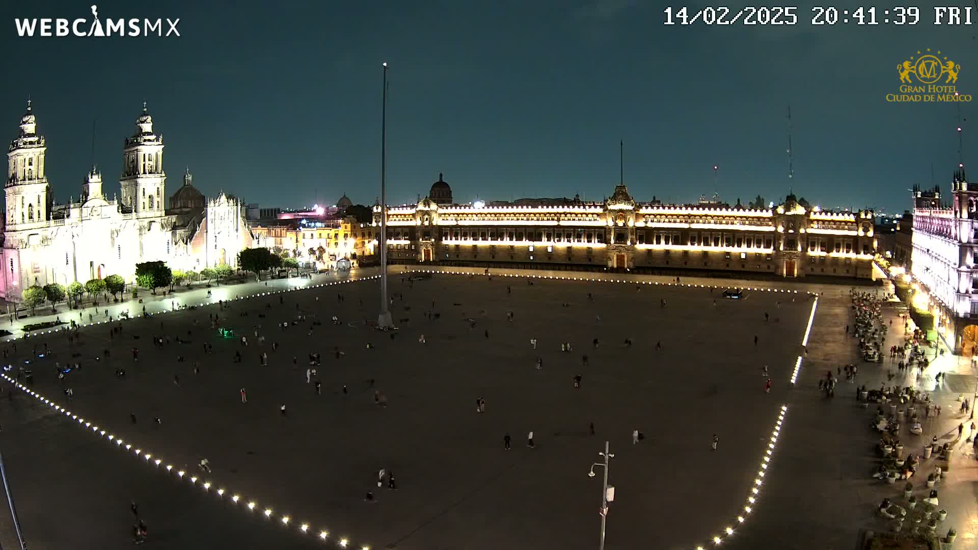 A large, mostly empty plaza at night is illuminated, with a cathedral and government buildings visible in the background under a dark sky.