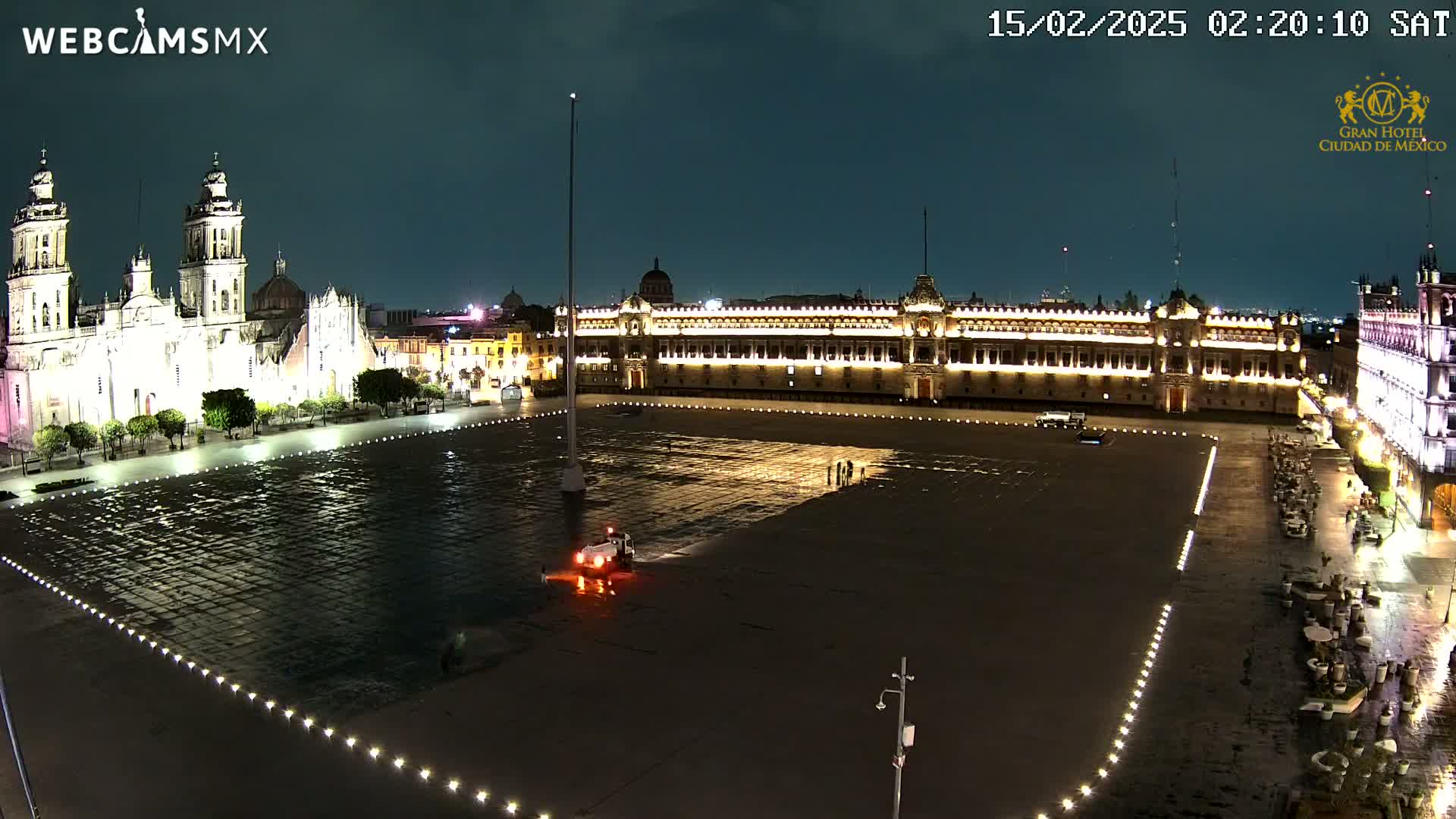 A large, dark plaza at night, illuminated by lights outlining its edges and buildings, is wet from recent rain, with a cathedral and government buildings visible in the background.