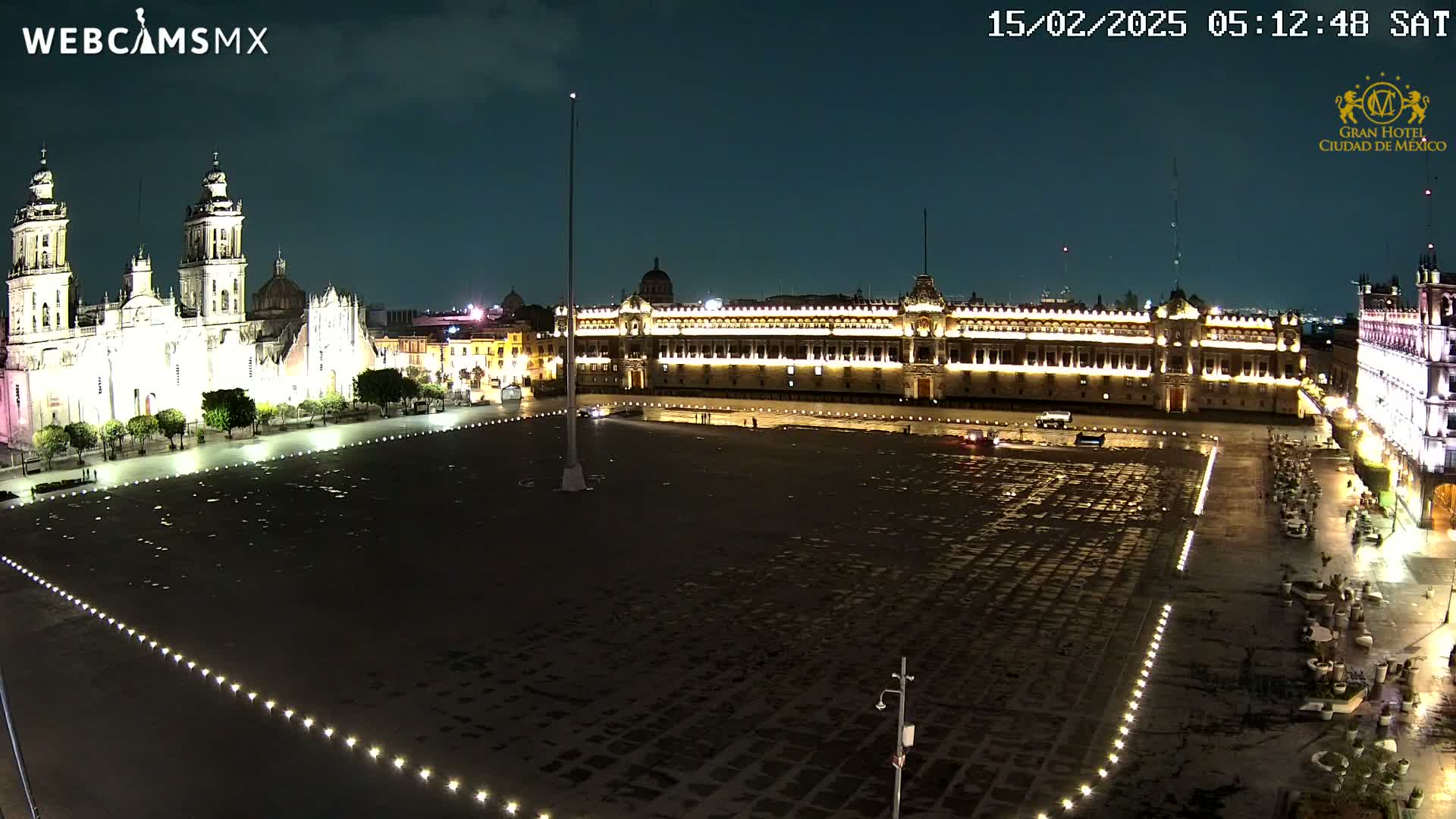 A large, empty plaza at night is lit by small lights along its edges, with a brightly lit cathedral and government building visible on either side.