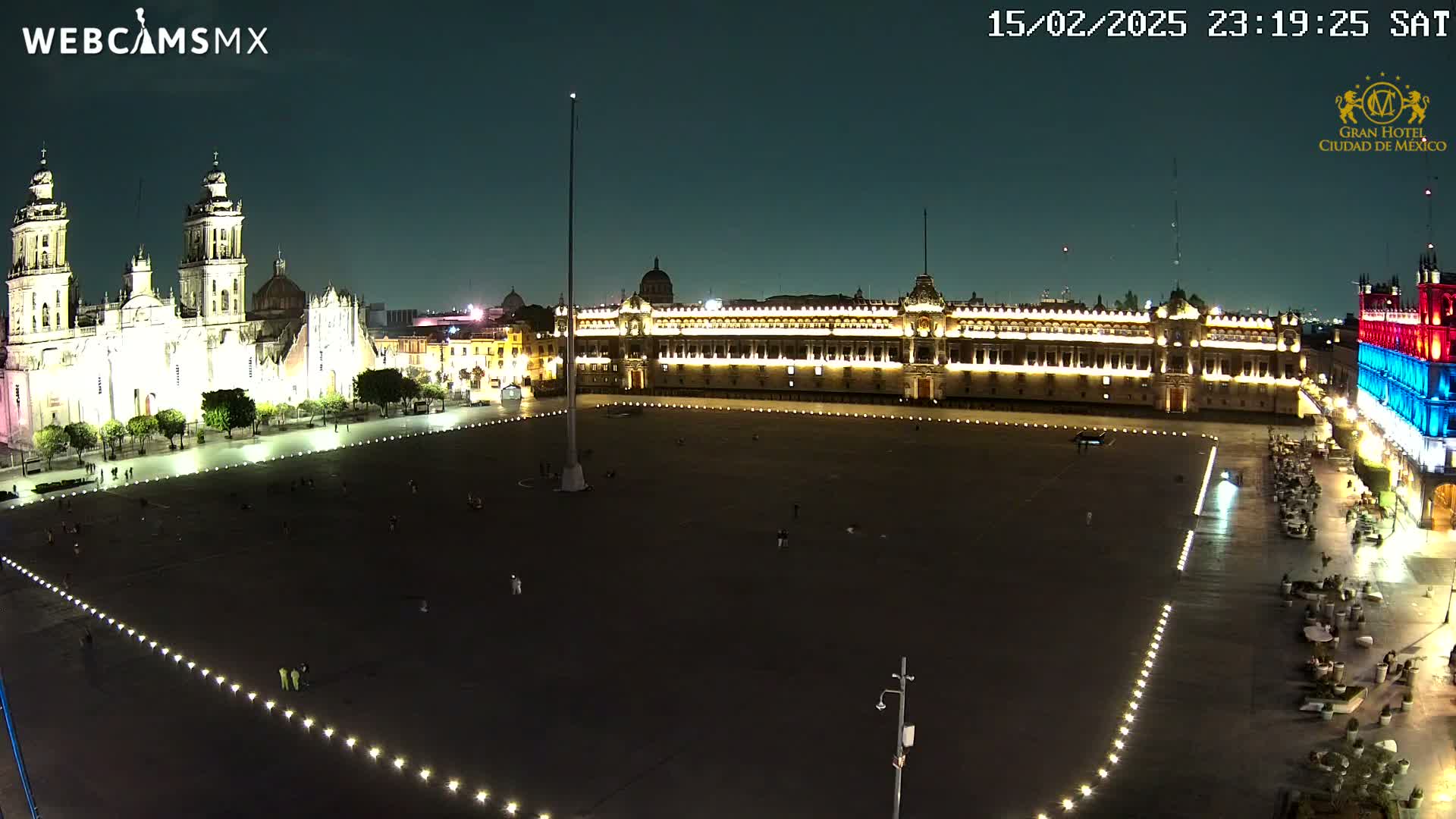 A large, mostly empty plaza at night is illuminated, with a cathedral and government building visible on opposite sides.