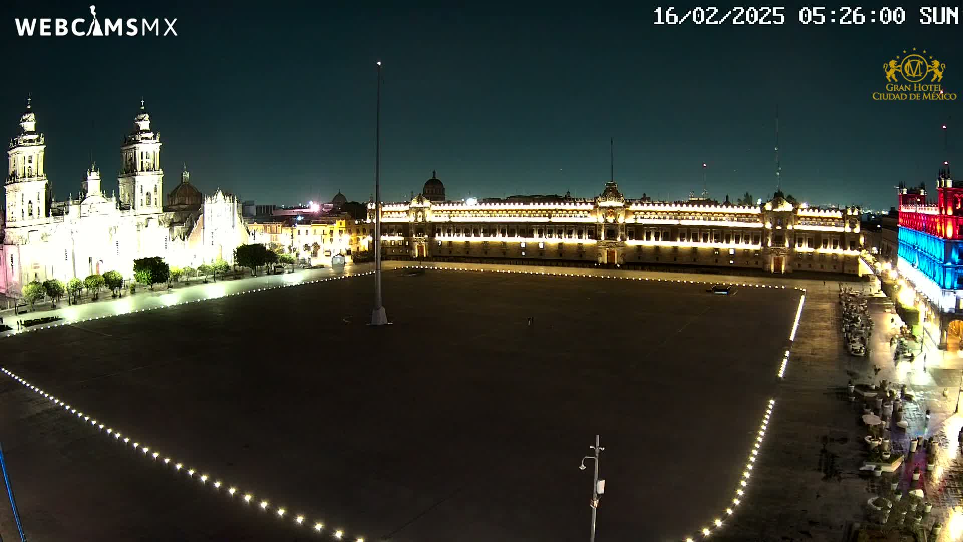 A large, mostly empty plaza at night is illuminated, bordered by a brightly lit cathedral and government building.
