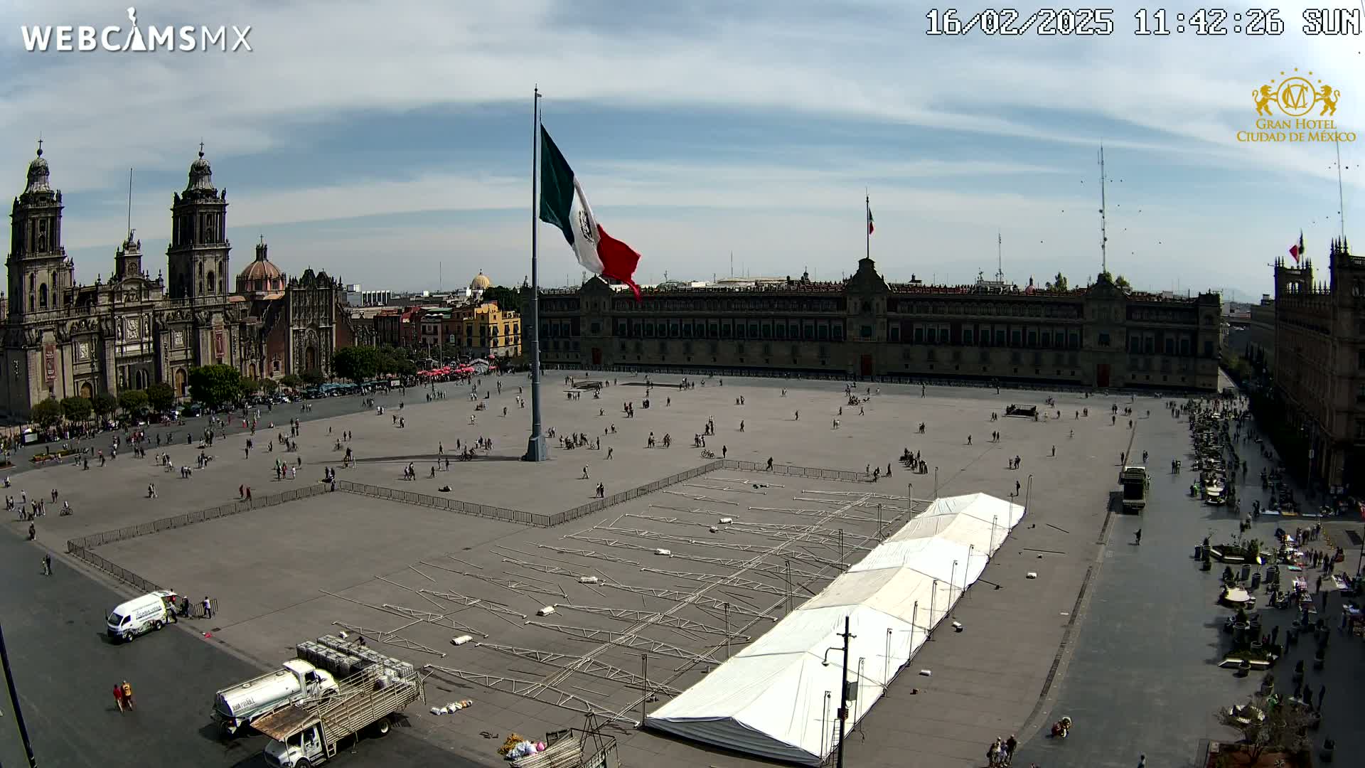 A large plaza with a partially erected tent, a Mexican flagpole, a cathedral, and a government building is seen under a mostly sunny sky.