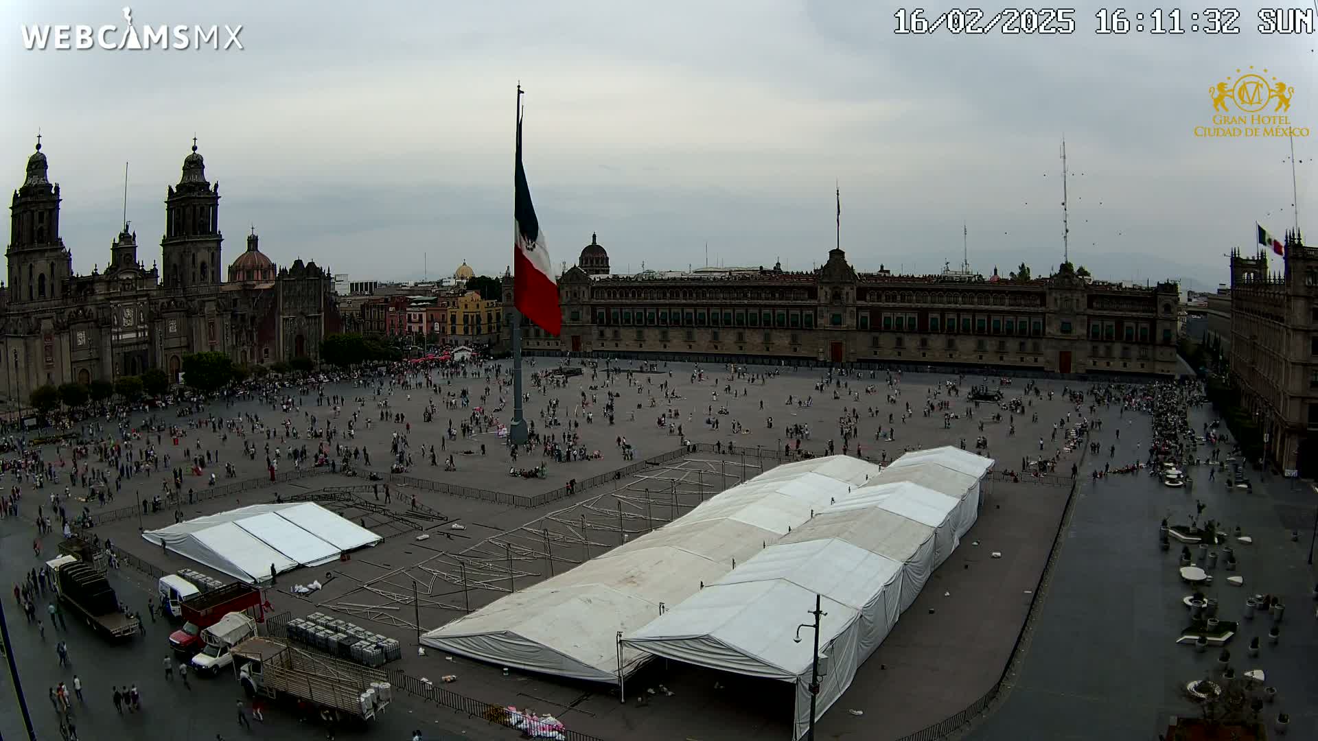 A large plaza with a crowd of people, a cathedral, and government buildings under an overcast sky, with several large white tents set up in the plaza.