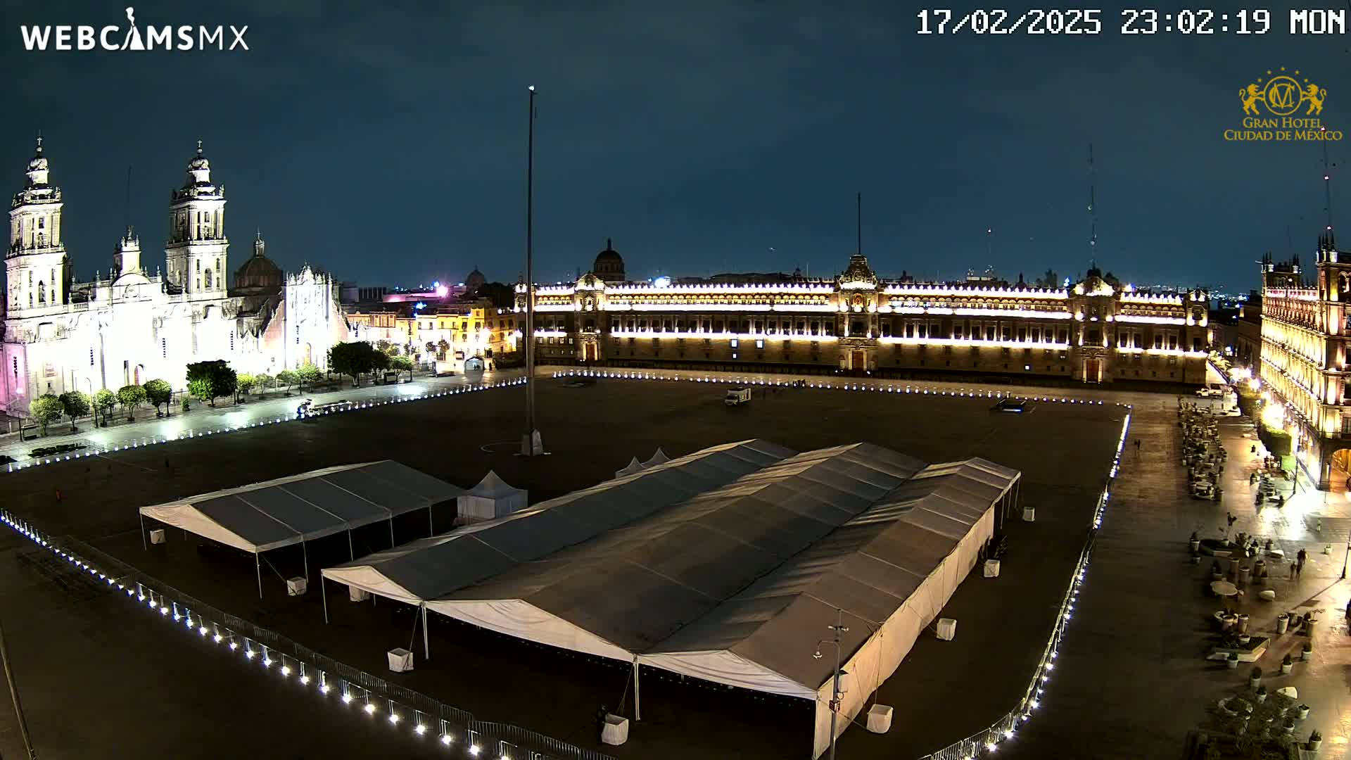 A nighttime, aerial view of a large plaza with several tents, a cathedral, and a long, illuminated building.