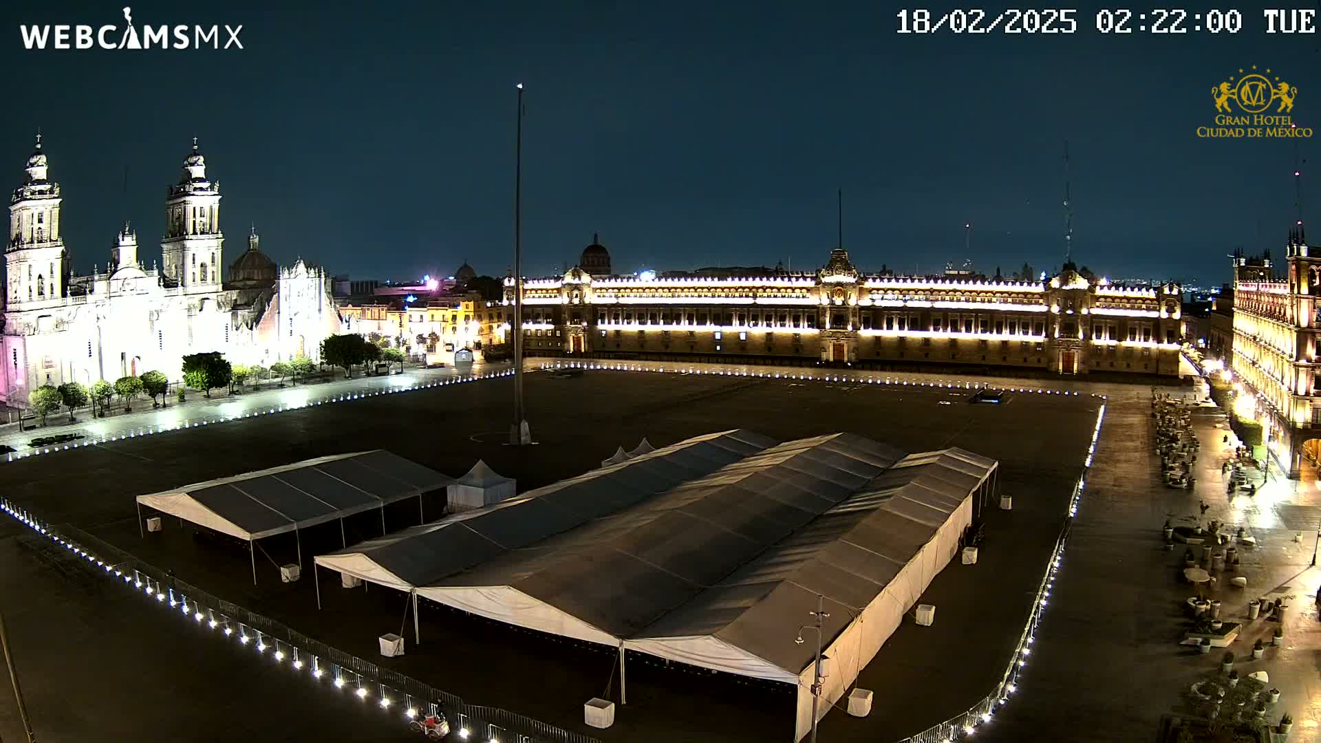 A nighttime view of a large plaza, featuring a brightly lit cathedral and palace, with several large tents set up in the center of the square under a clear, dark sky.