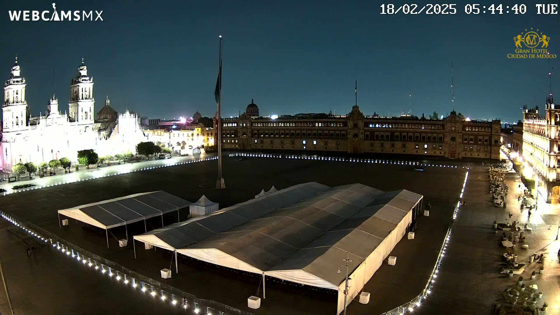 A nighttime, illuminated view of a large plaza with several tents set up, flanked by a cathedral and a long building.