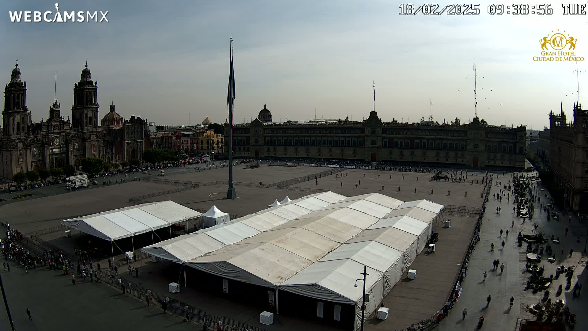 A large plaza with several white tents, a cathedral, and a long building, under a mostly cloudy sky.