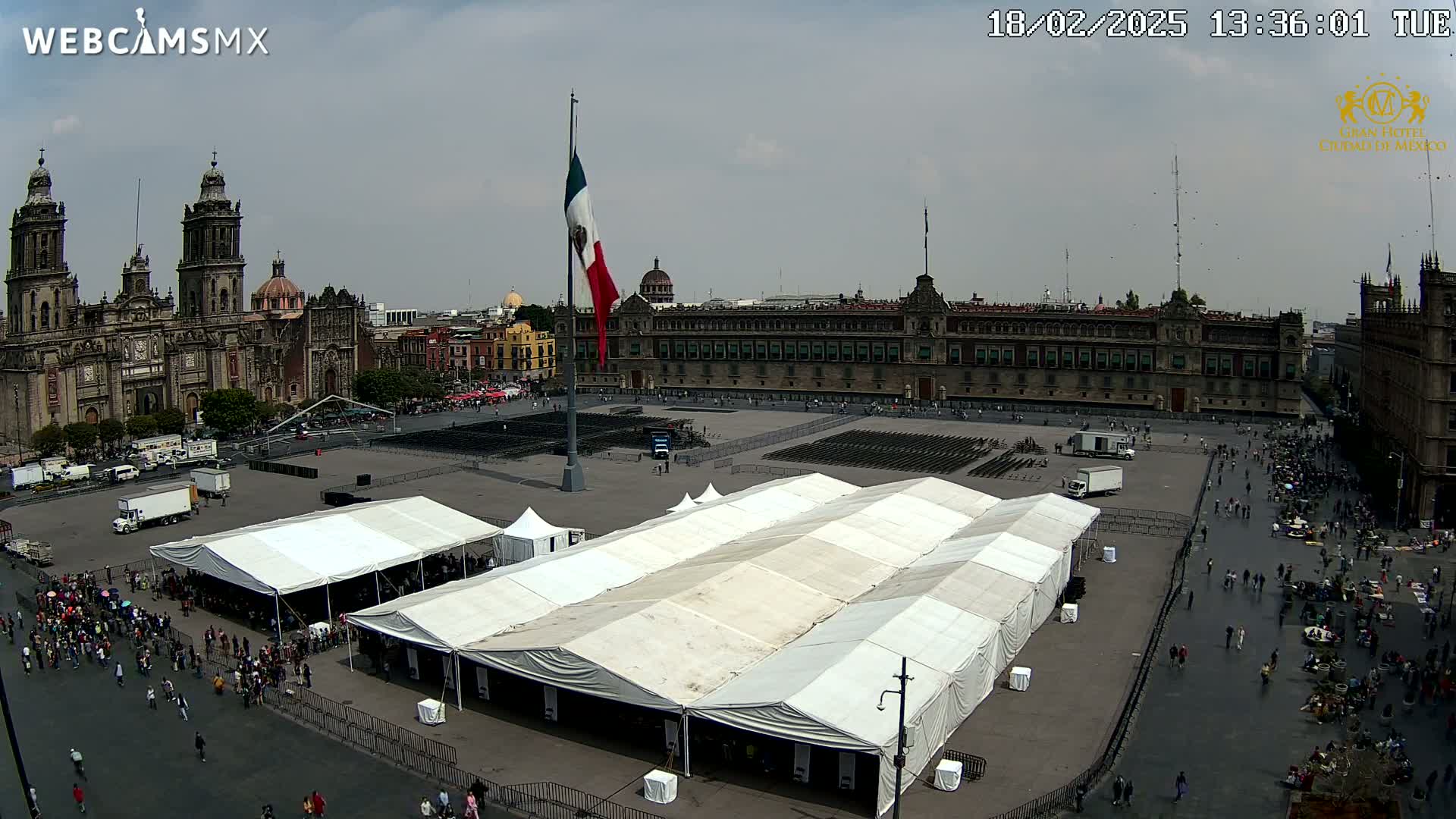A large plaza with several white tents, a Mexican flag on a tall pole, a cathedral, and a large building in the background under an overcast sky.