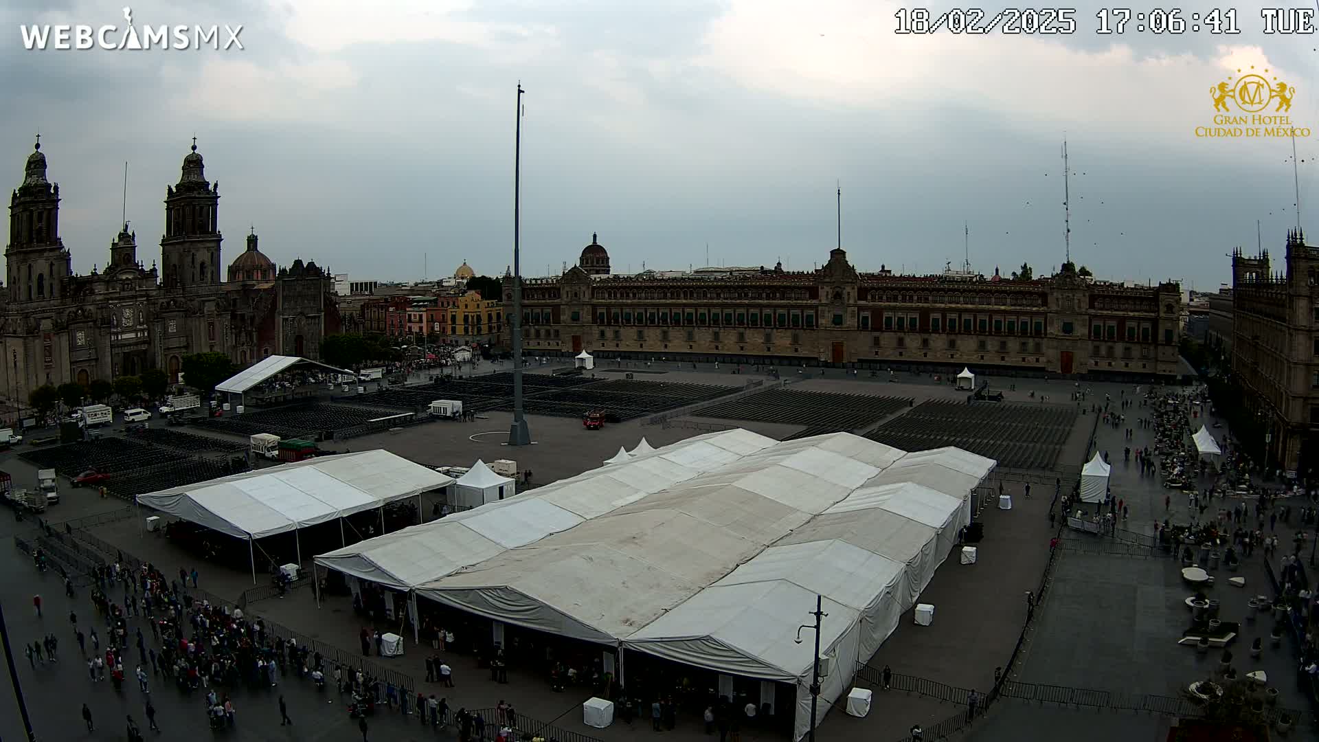 A large plaza with several white tents, many people, and a large building behind it under an overcast sky.