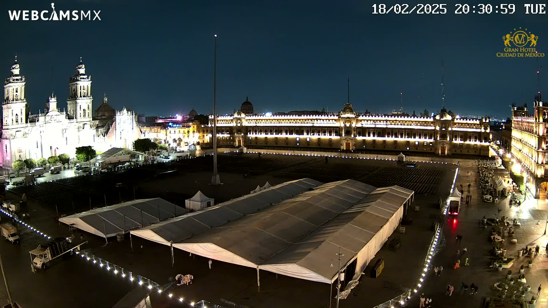 A large, illuminated plaza at night, featuring a cathedral and government buildings, with several large tents set up in the square.