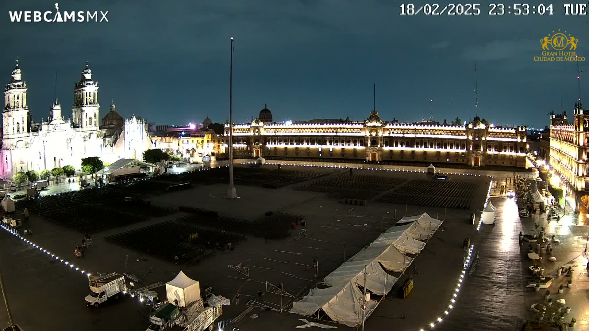 A large, mostly empty plaza at night, illuminated by lights, features a cathedral and government building, with several tents and a few vehicles visible.