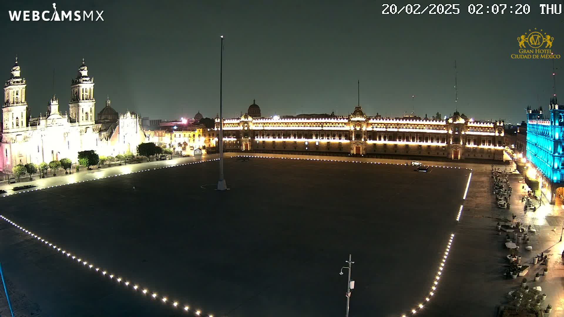 A large, dark plaza at night is illuminated by lights lining its edges, with a brightly lit cathedral and palace visible on opposite sides.