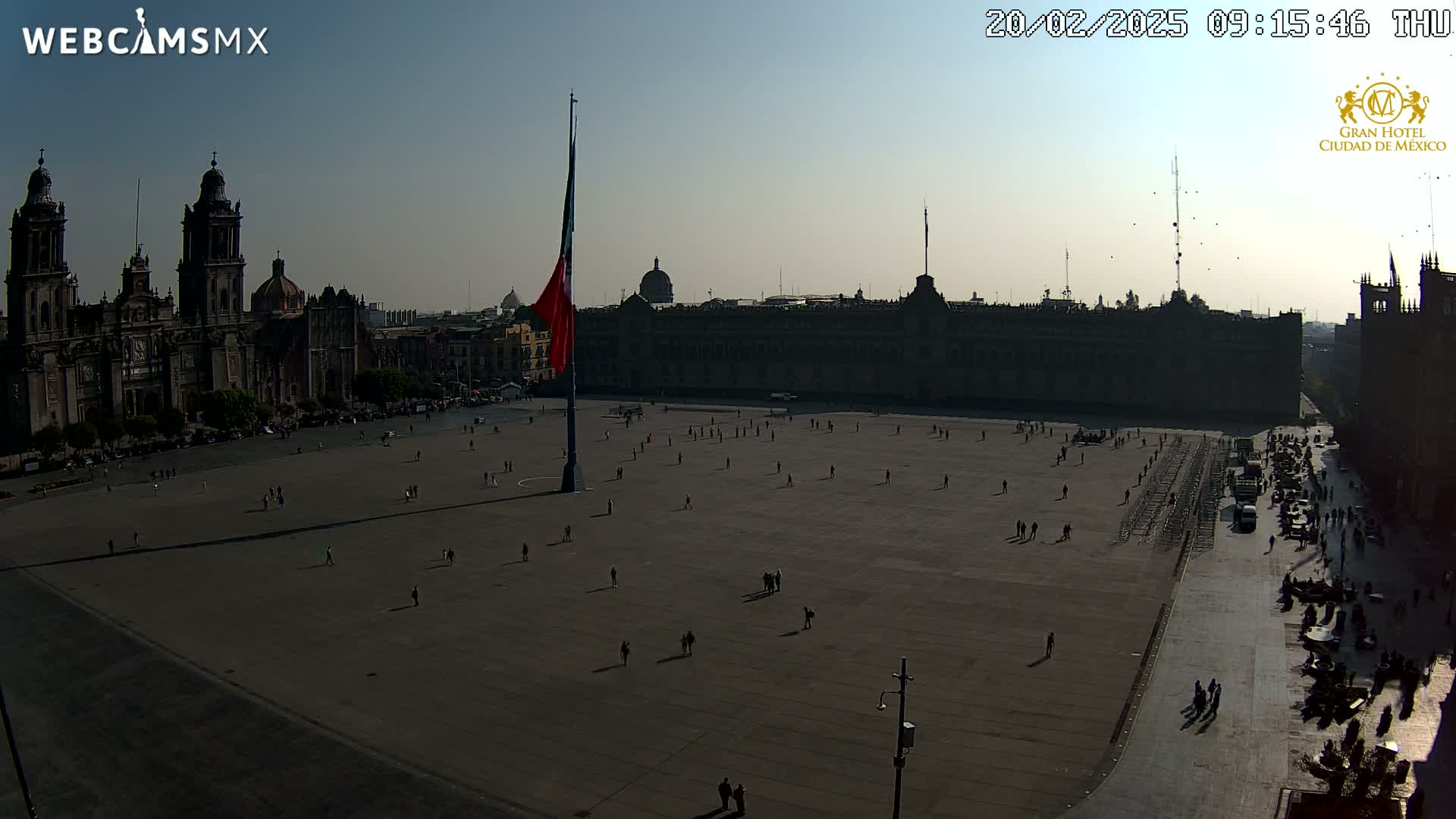 A large plaza with a few people sparsely scattered across it, a large building and cathedral in the background under a clear, sunny sky.