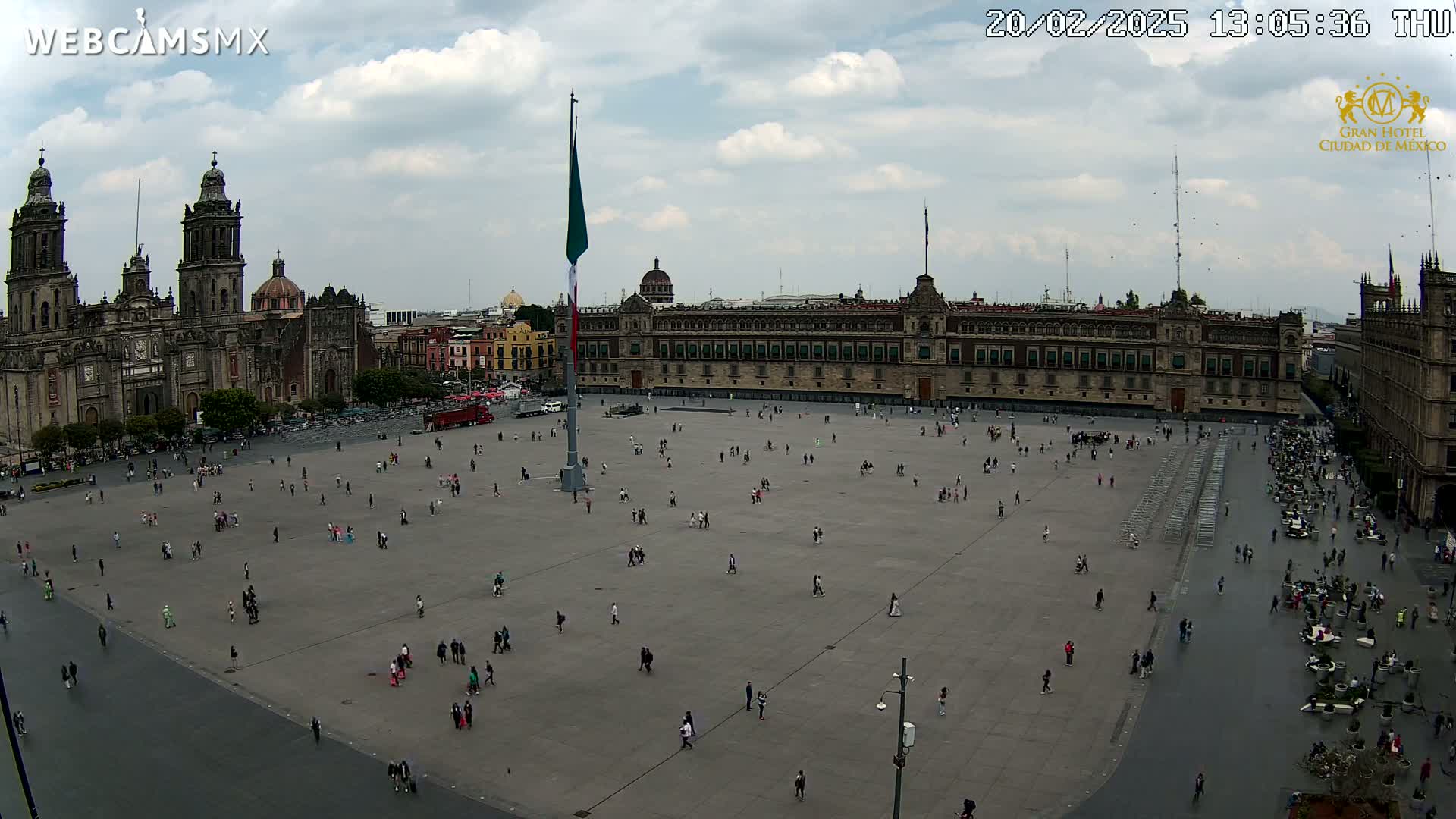 A large plaza with a sparsely populated crowd is viewed from above under a partly cloudy sky, with a cathedral and government buildings visible in the background.