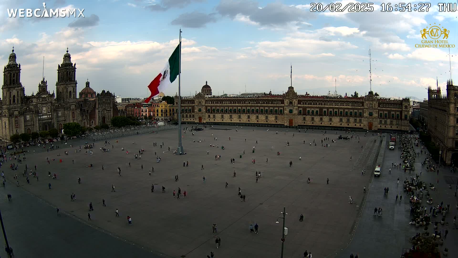 A large plaza with sparsely scattered people is seen, under partly cloudy skies, with a large Mexican flag flying in front of a grand building and a cathedral in the background.