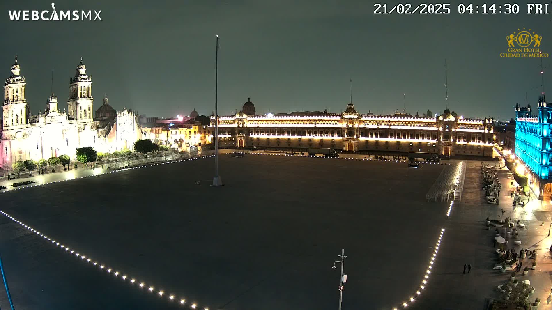 A large, dark plaza at night is illuminated by lights lining its edges, with a brightly lit cathedral and government building visible in the background under a clear night sky.