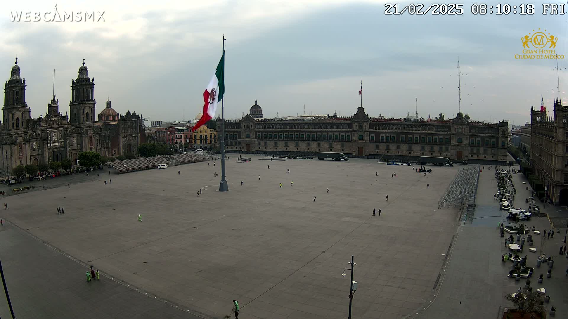 A large plaza with a Mexican flag at its center, surrounded by historical buildings under an overcast sky.