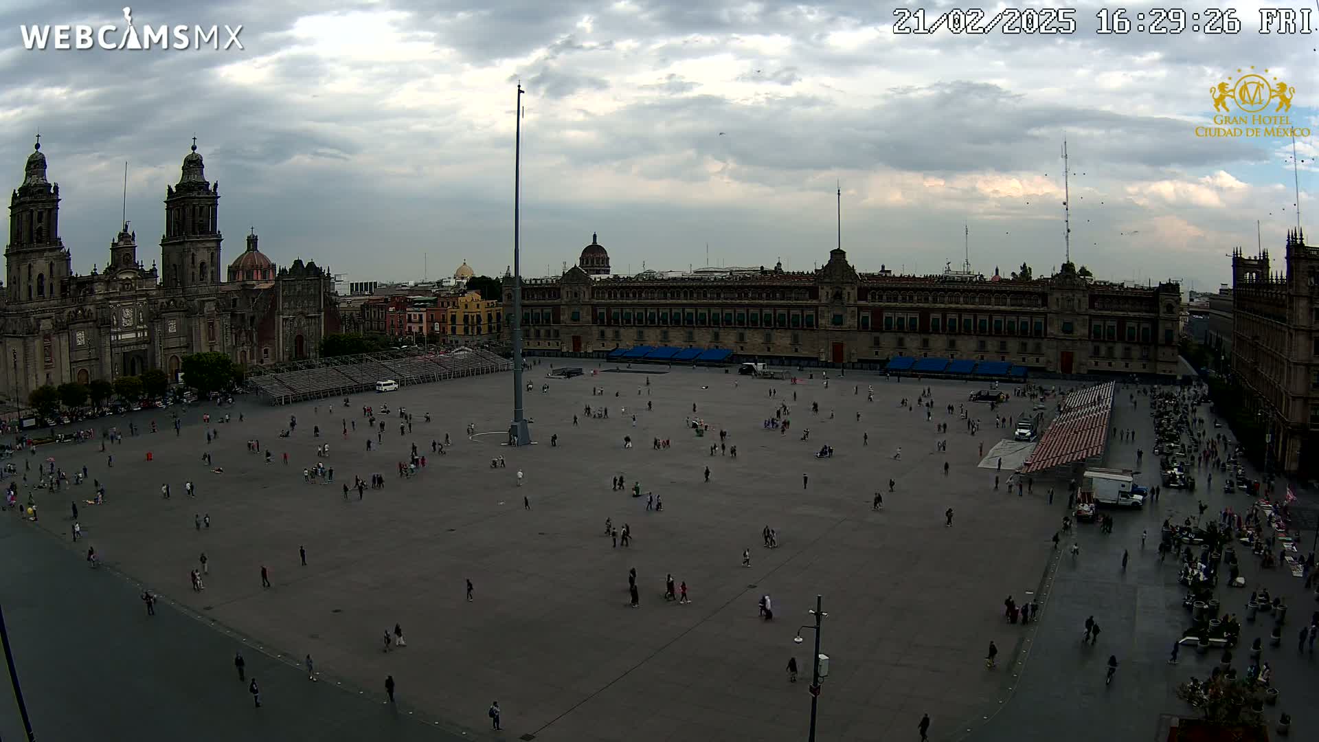 A large plaza with many people sparsely scattered across it is situated in front of a large, historic building and a cathedral under a cloudy sky.