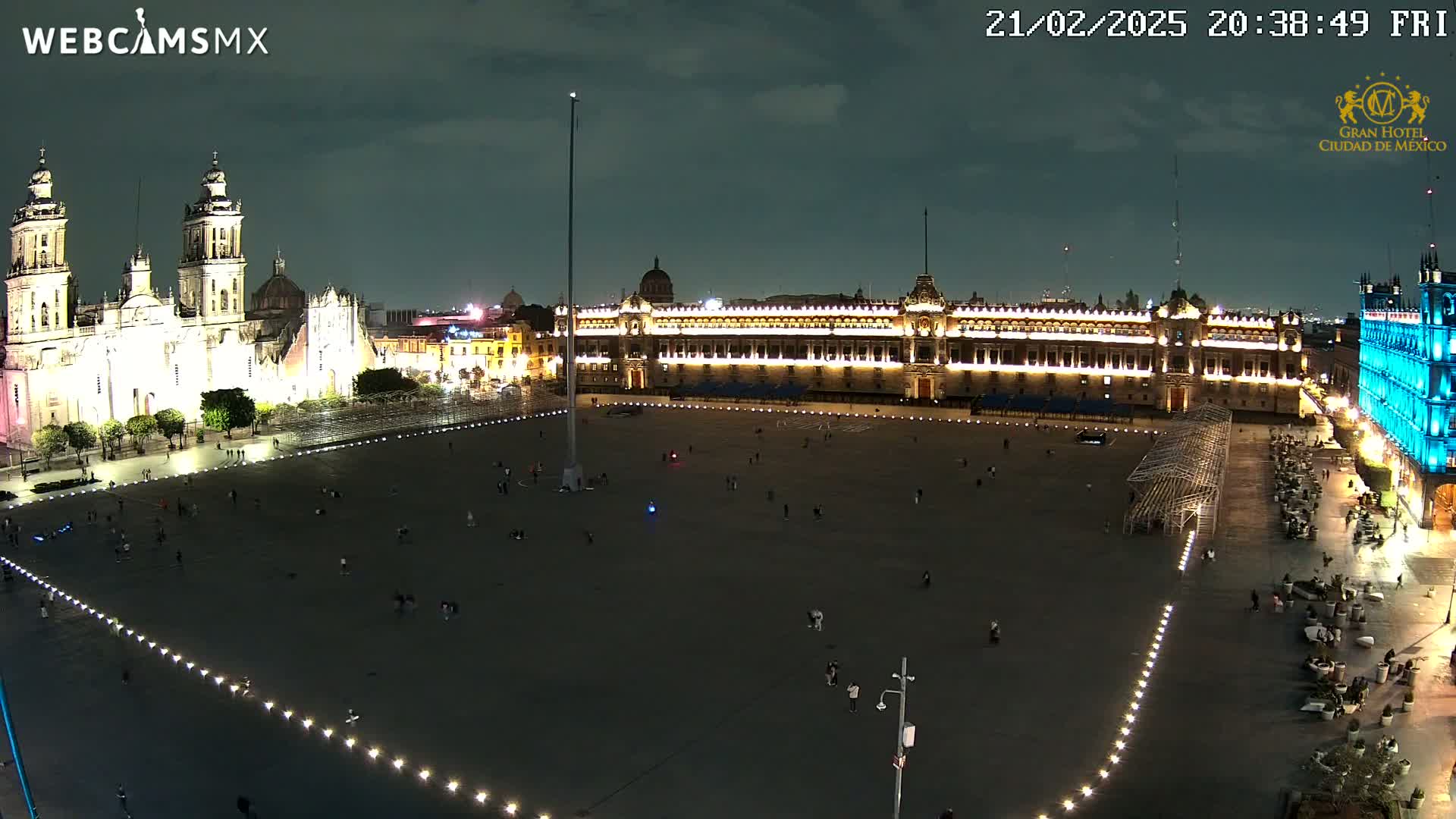 A large, mostly empty plaza at night is illuminated, with a brightly lit cathedral and palace visible in the background under a partly cloudy sky.
