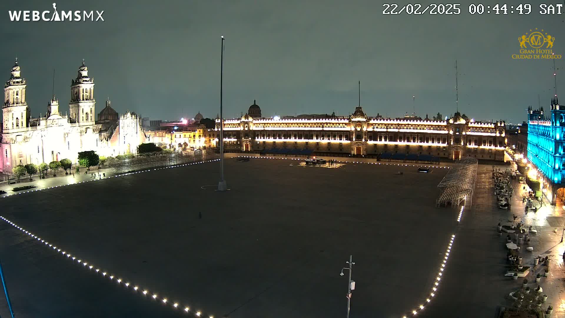 A large, dark plaza at night is illuminated by lights, with a brightly lit cathedral and palace visible in the background under a dark, possibly overcast sky.